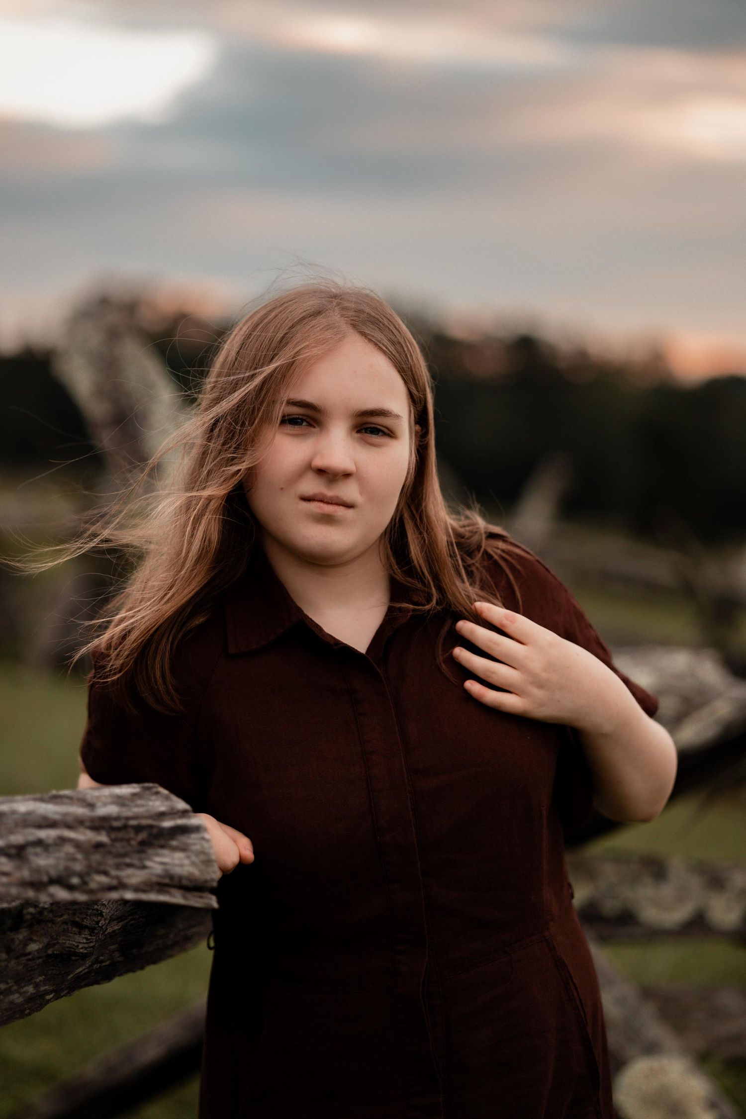 Young woman in dark dress leaning on rustic fence at sunset with wind-blown hair in outdoor portrait.