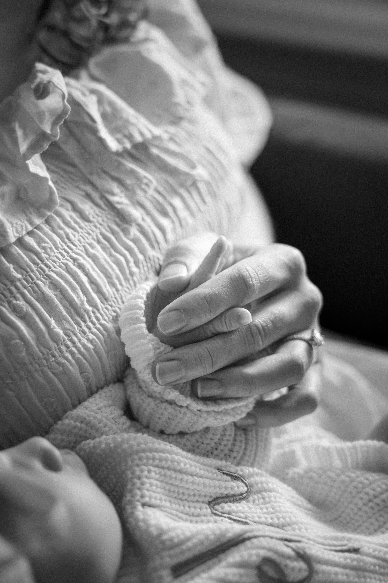 Black and white photo of a mother holding her newborn baby's tiny hand, wearing a smocked dress and ring.