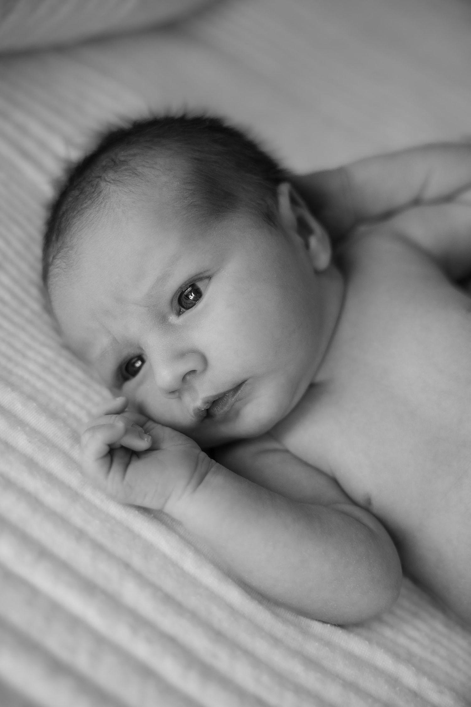 Black and white photo of a newborn baby lying on a soft blanket, gazing upward with tiny hands near face.