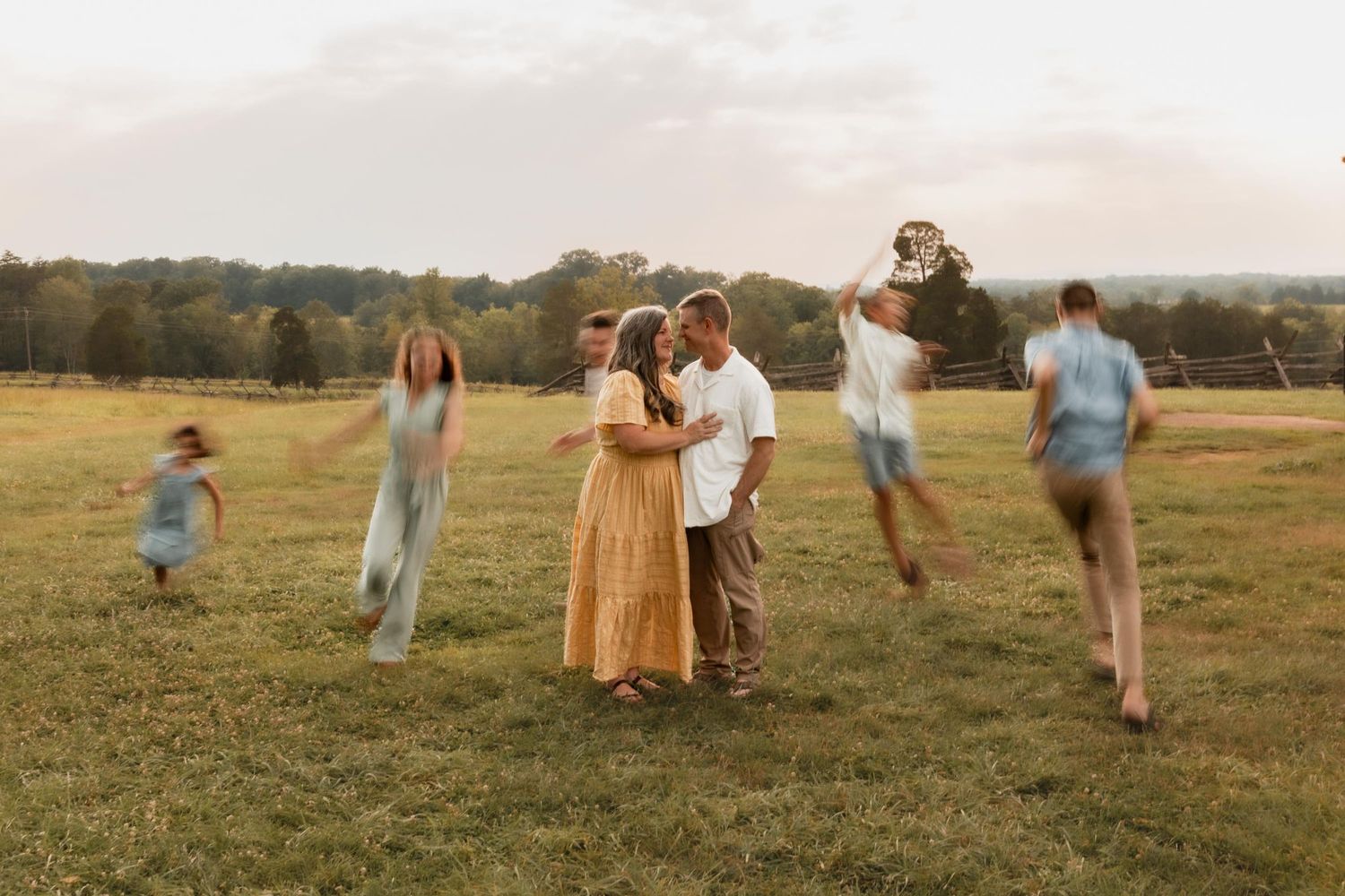 Couple shares tender moment in golden field while children play around them during family portrait session.