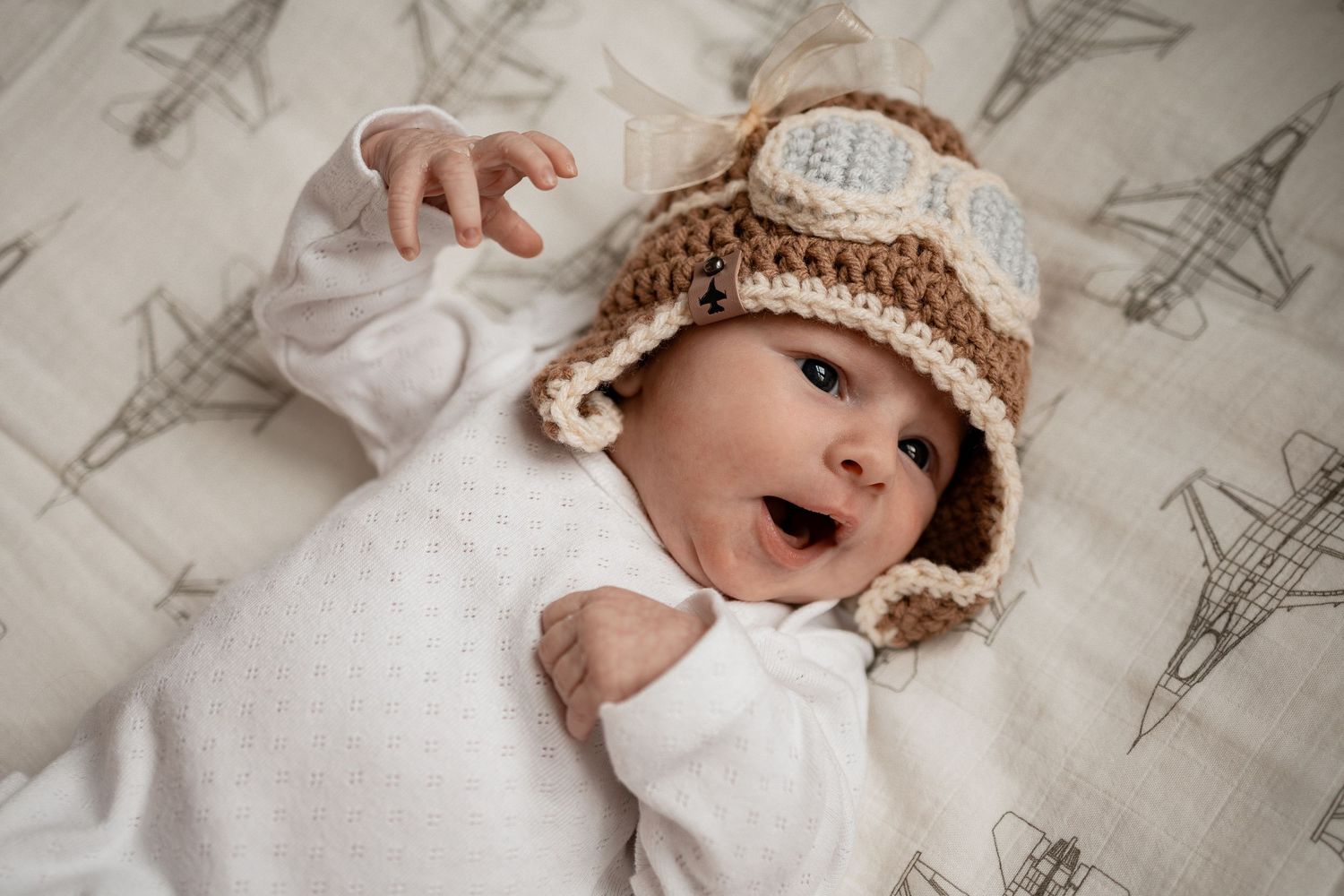 Baby wearing brown knitted aviator hat lies on fabric with Eiffel Tower print background.