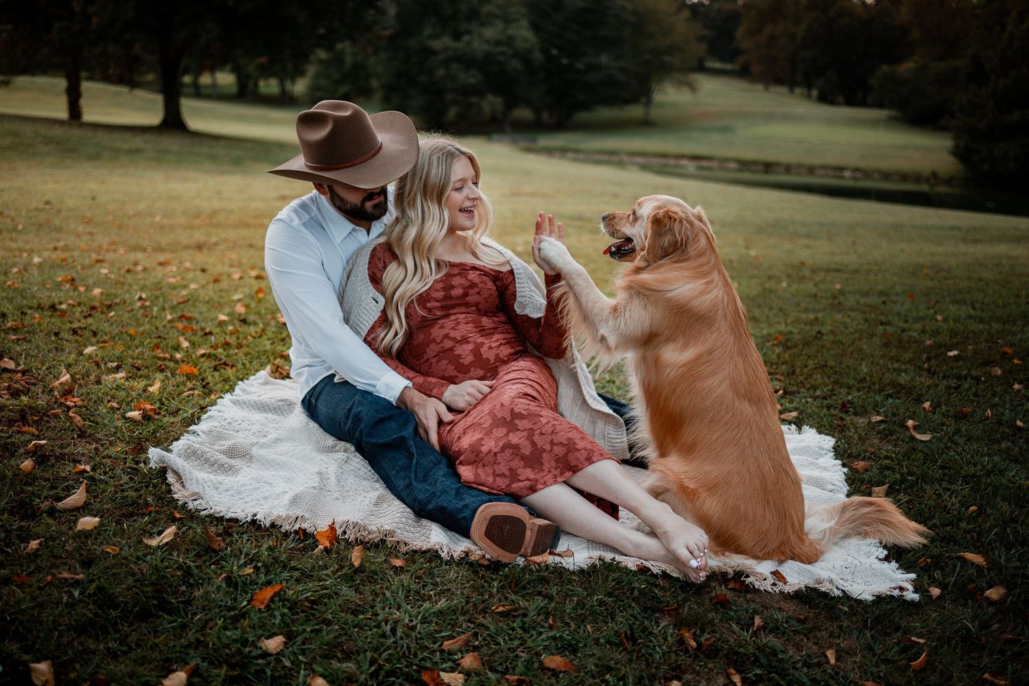 Maternity picnic scene with couple and dog relaxing on blanket in autumn park setting.