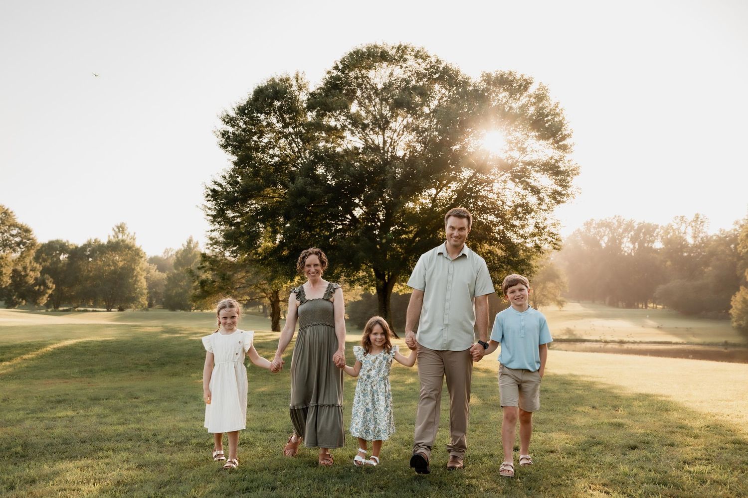 Family of five holding hands in a sunny park with golden hour light filtering through large green trees.