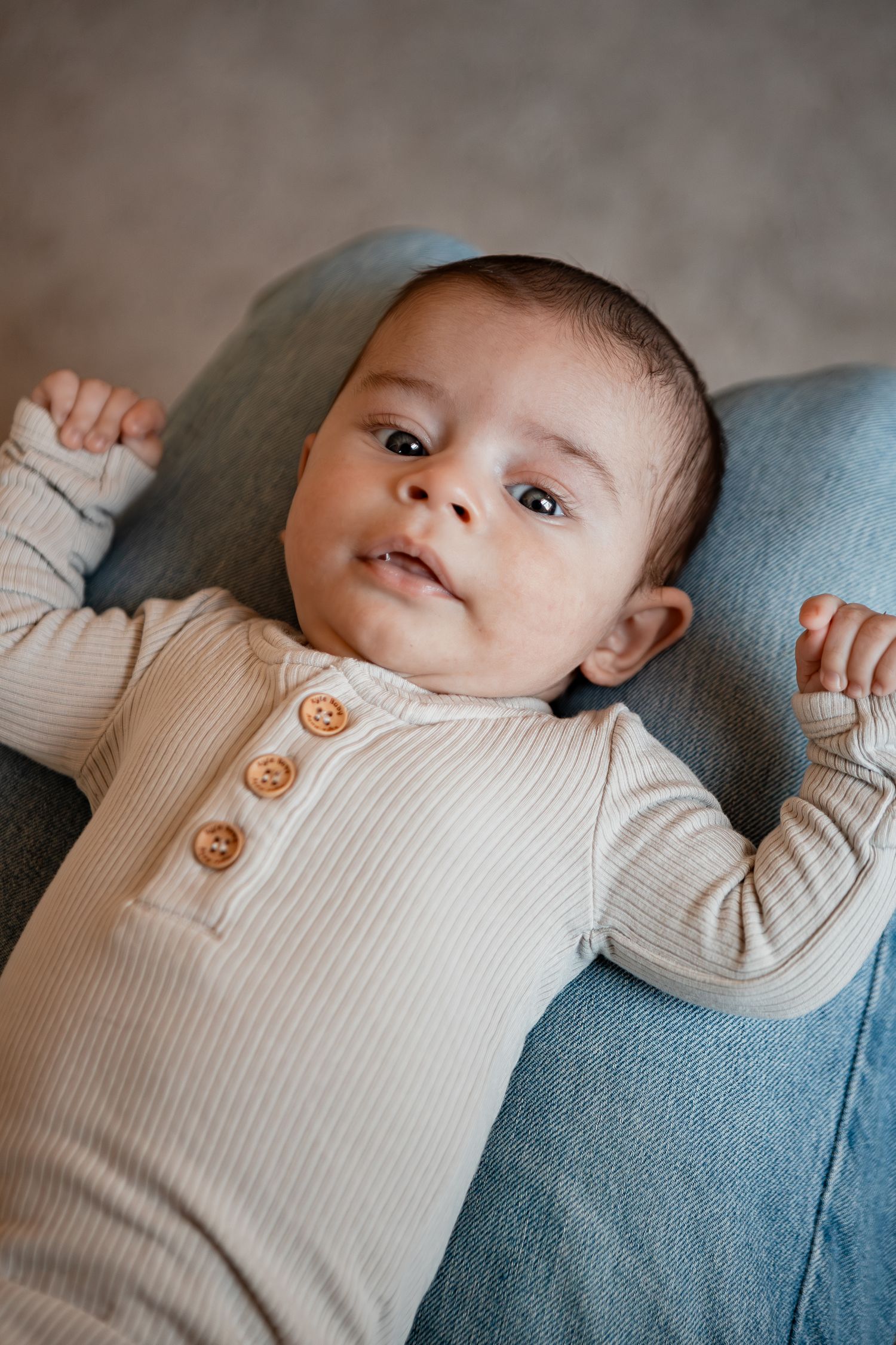A baby in a cream colored ribbed outfit lies on a blue cushion while looking at the camera.