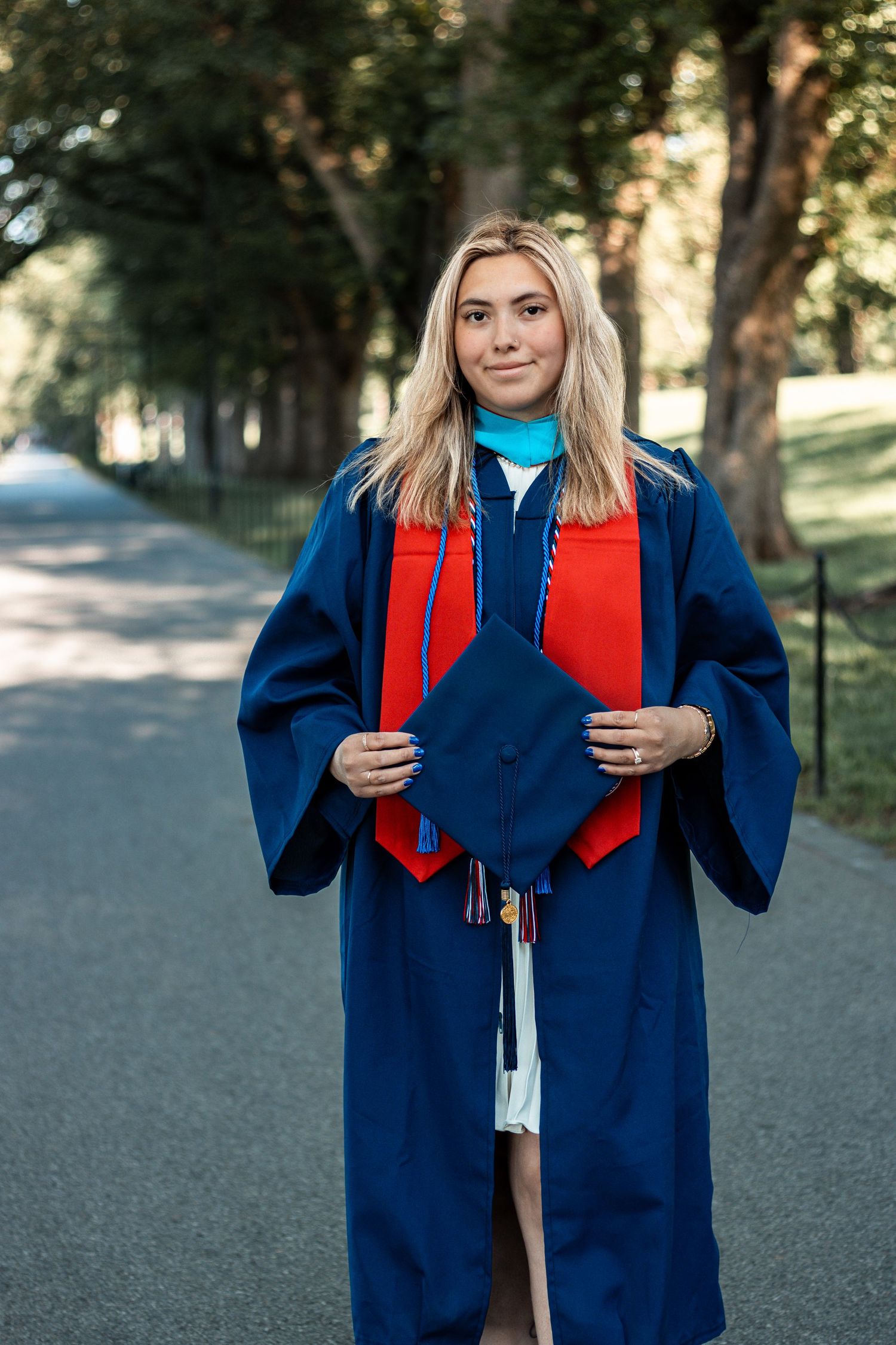 Graduate wearing blue graduation gown with red stole standing in an outdoor park setting.