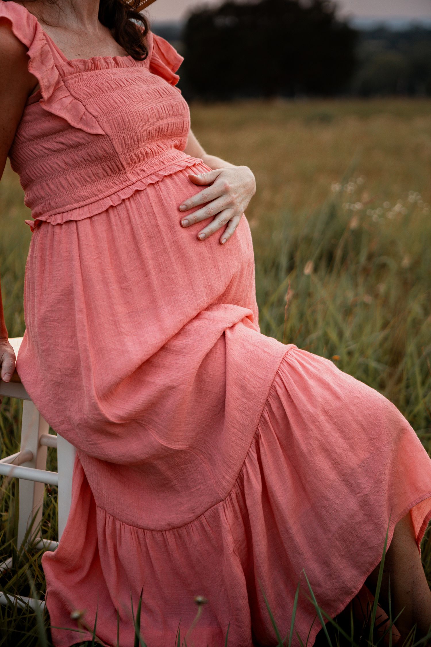 A person in a flowing coral dress sits gracefully in a summer field at dusk.