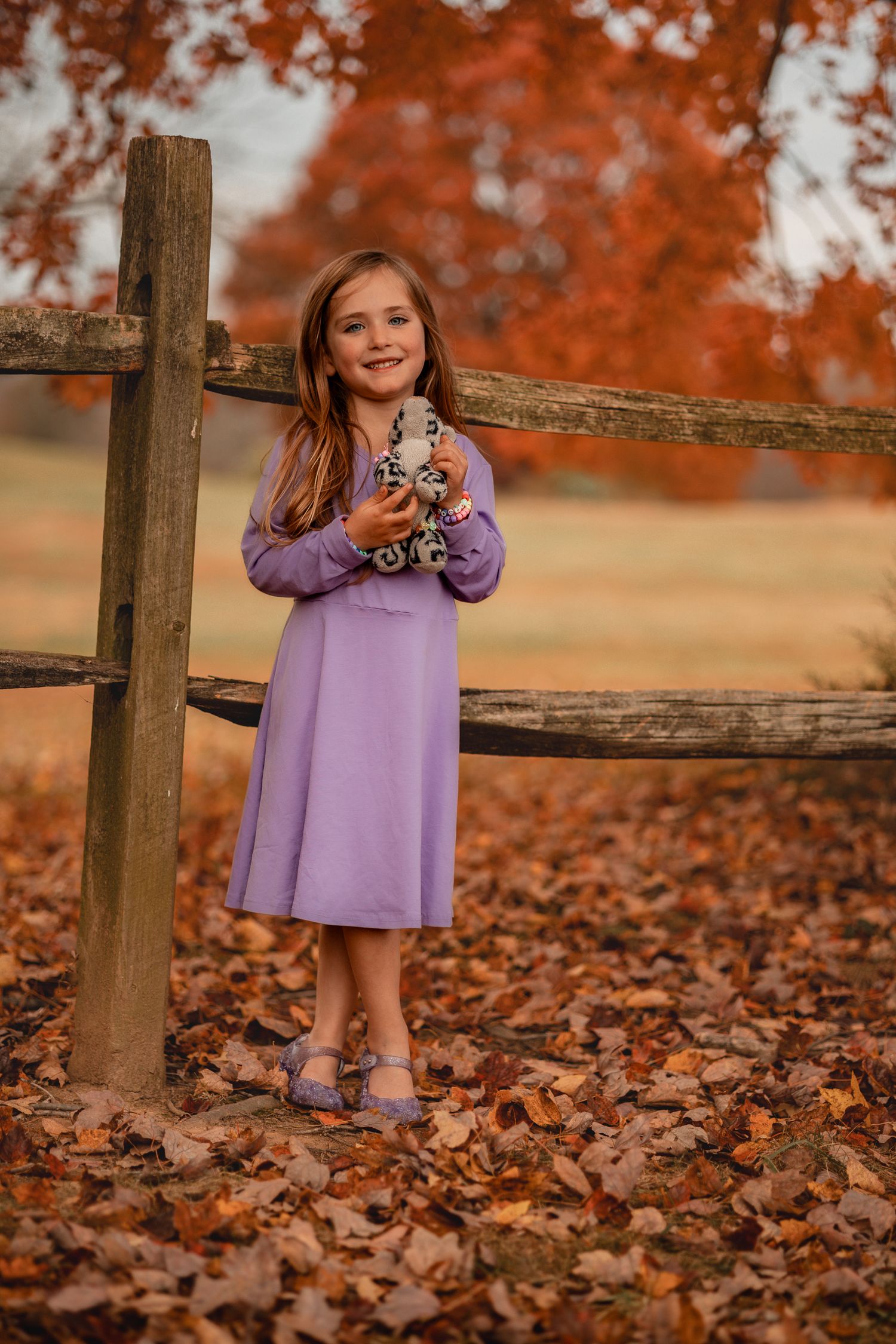 A child in a purple dress holds a toy while standing by a rustic wooden fence during autumn.
