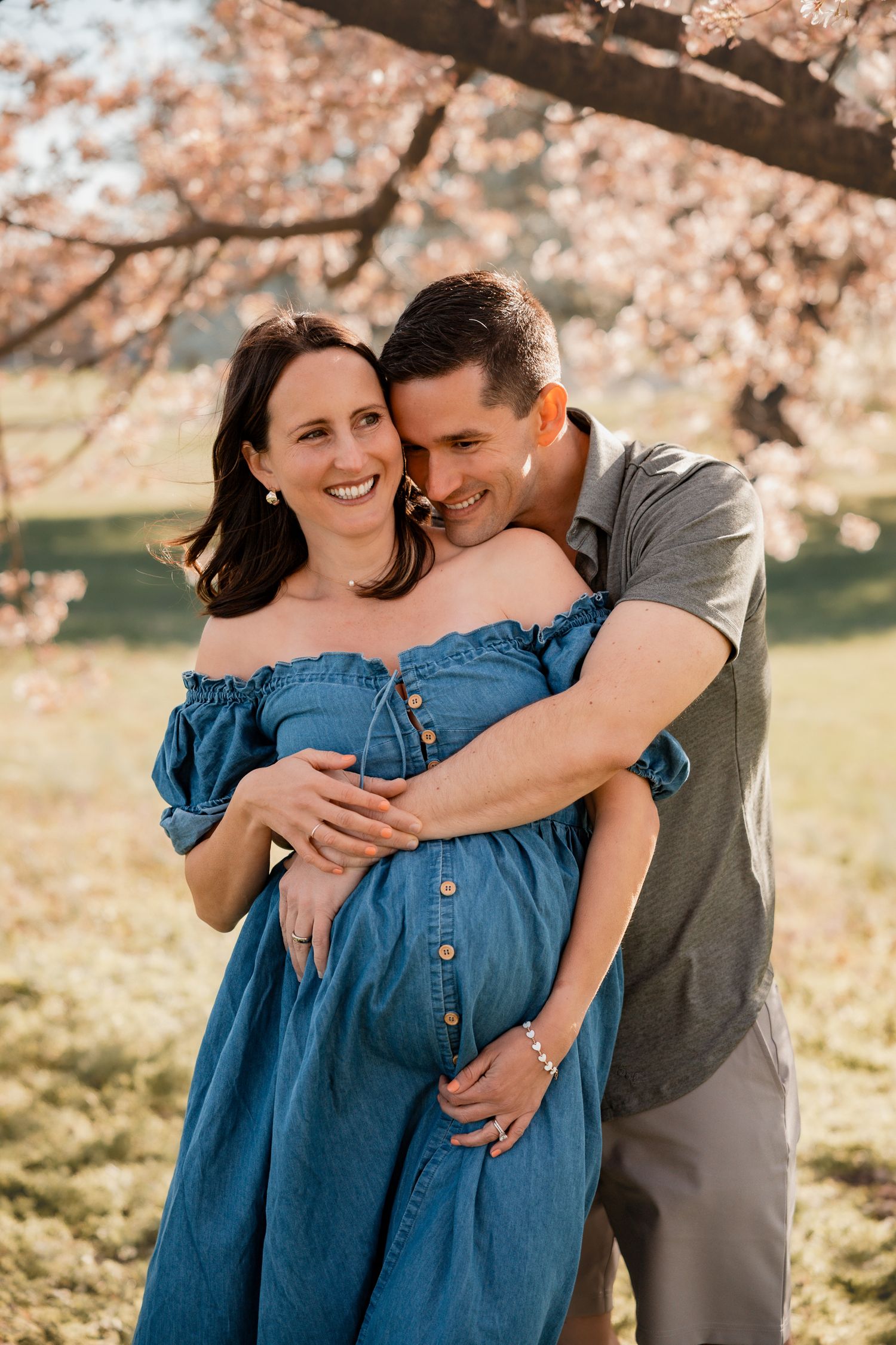 Couple embracing outdoors under cherry blossom trees, pregnant woman in blue off-shoulder dress smiling joyfully.