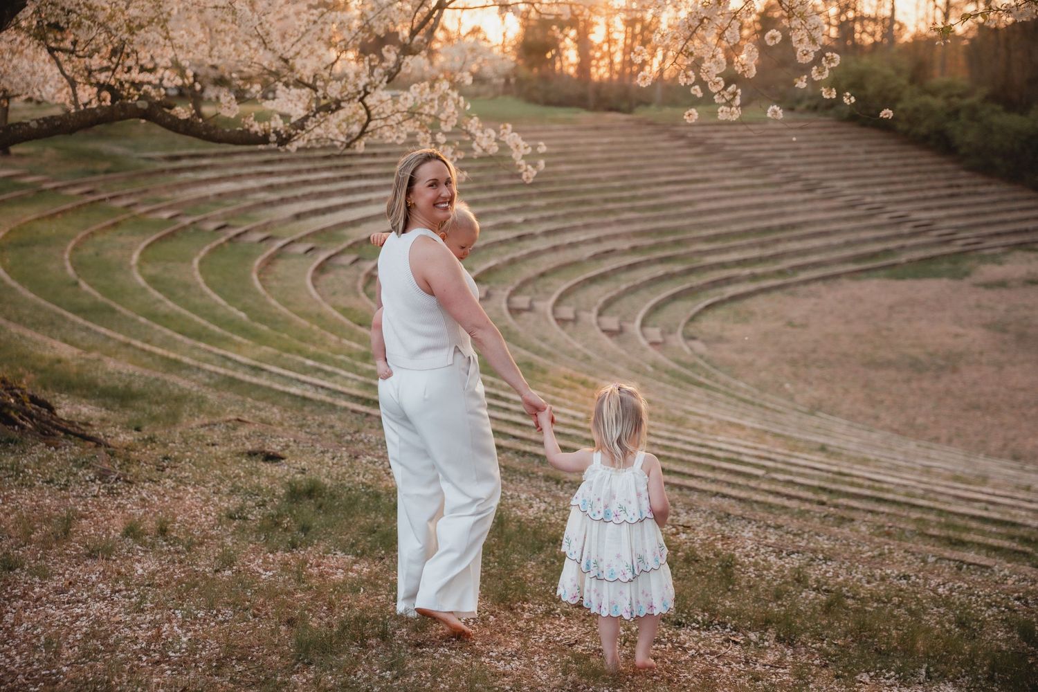 Mother and daughter in white outfits holding hands at sunset near a blooming tree amphitheater.