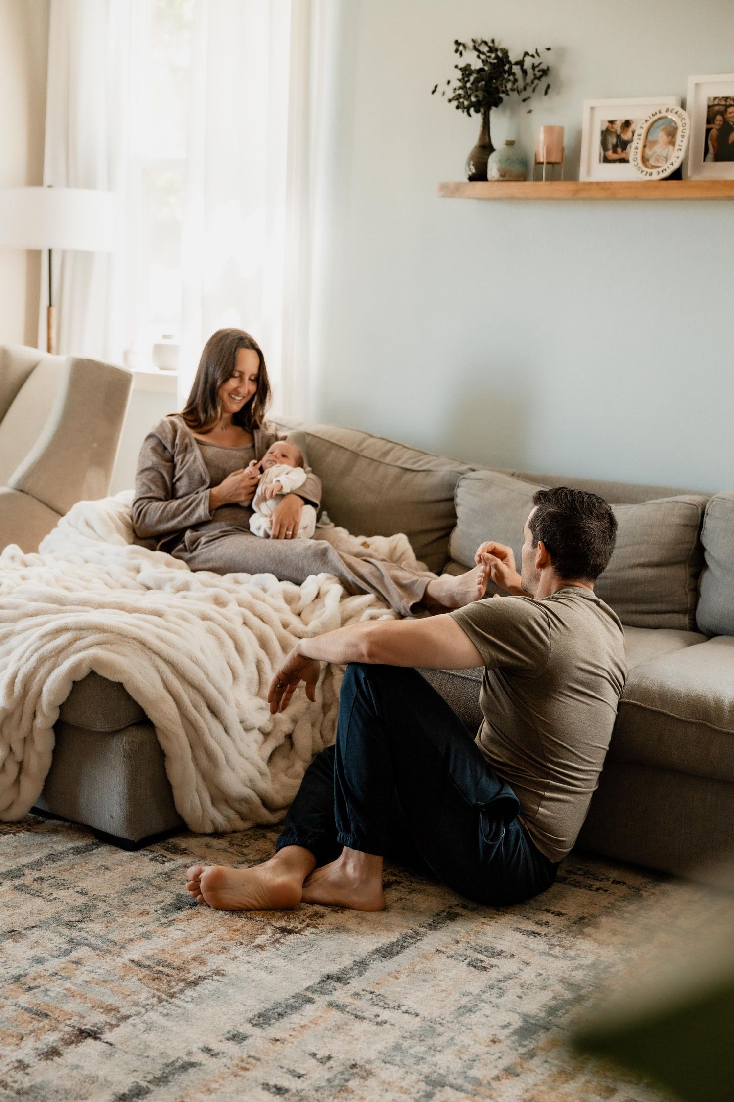 A young family sits together in a cozy living room, with a mother holding a newborn baby on the sofa.