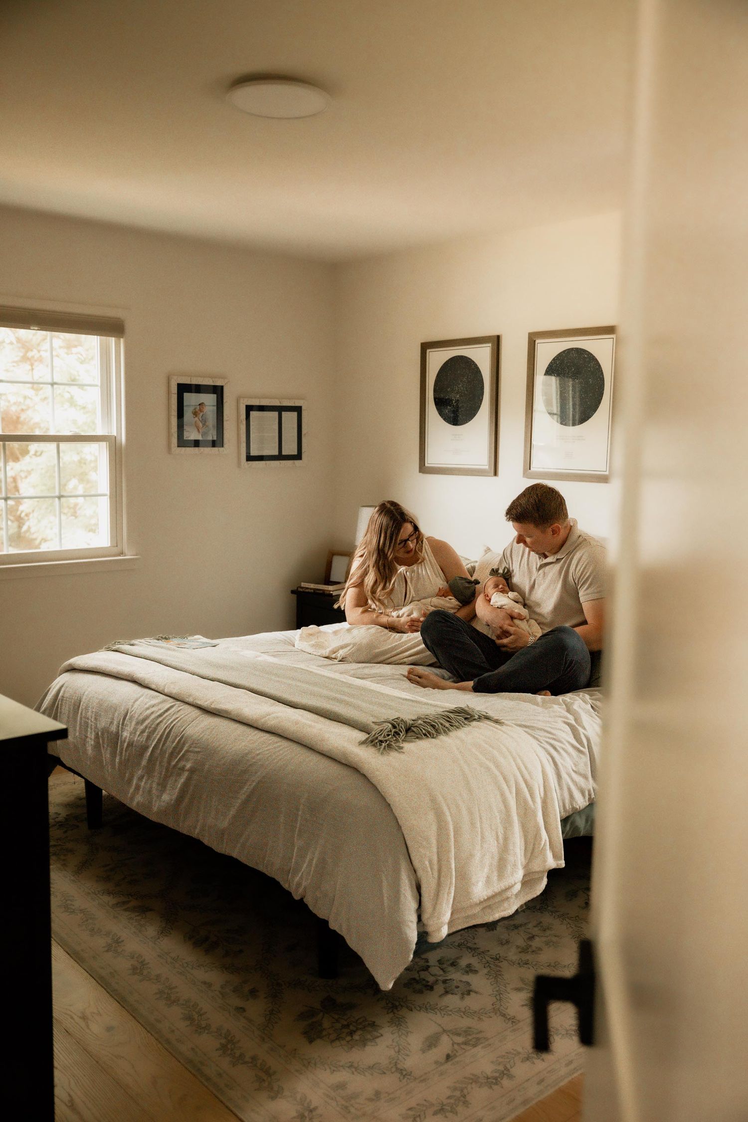 A couple sits on a bed with a newborn baby in a warmly lit bedroom with framed art on the walls.