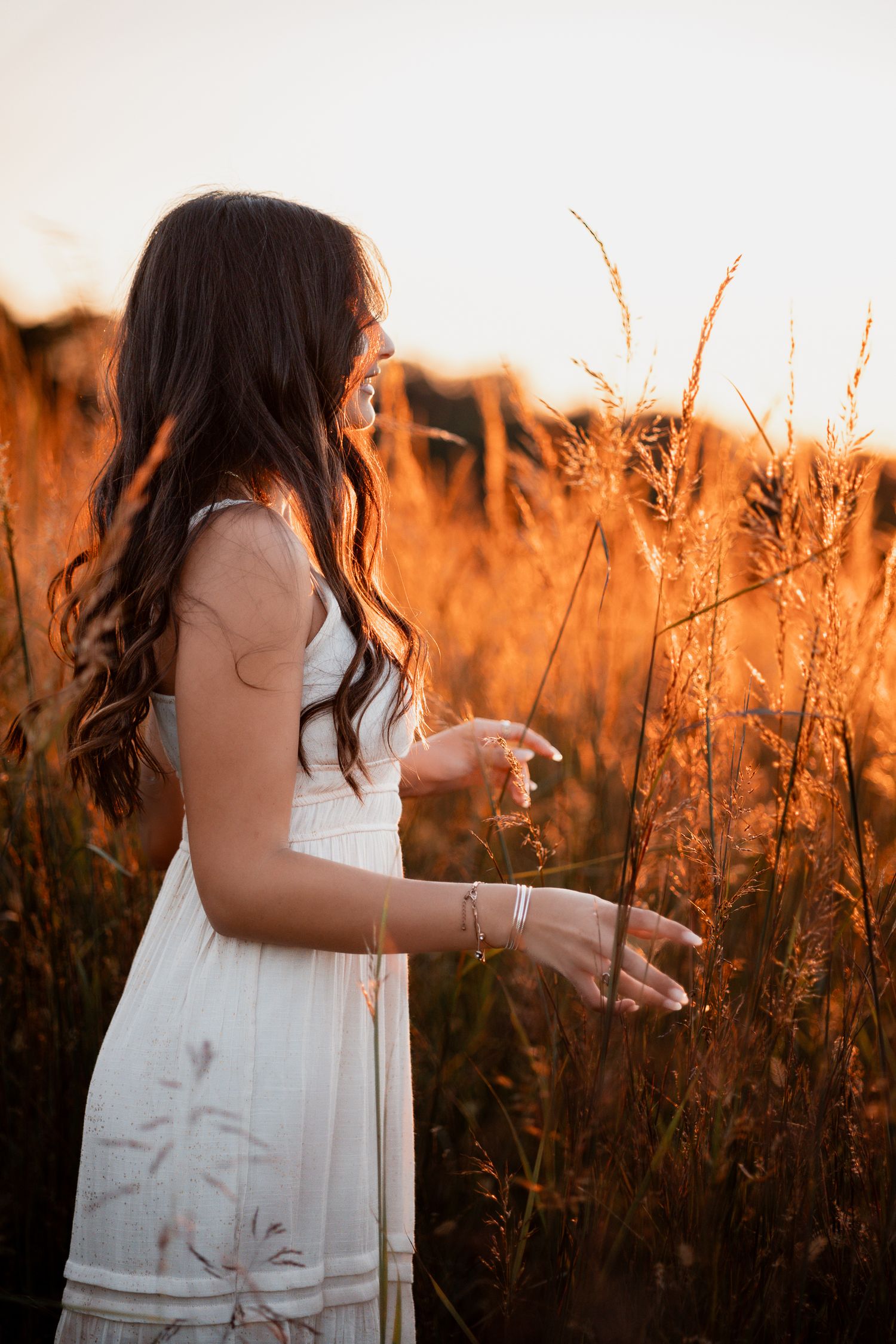 A woman in a white dress stands among tall golden grasses during sunset in a dreamy summer field.