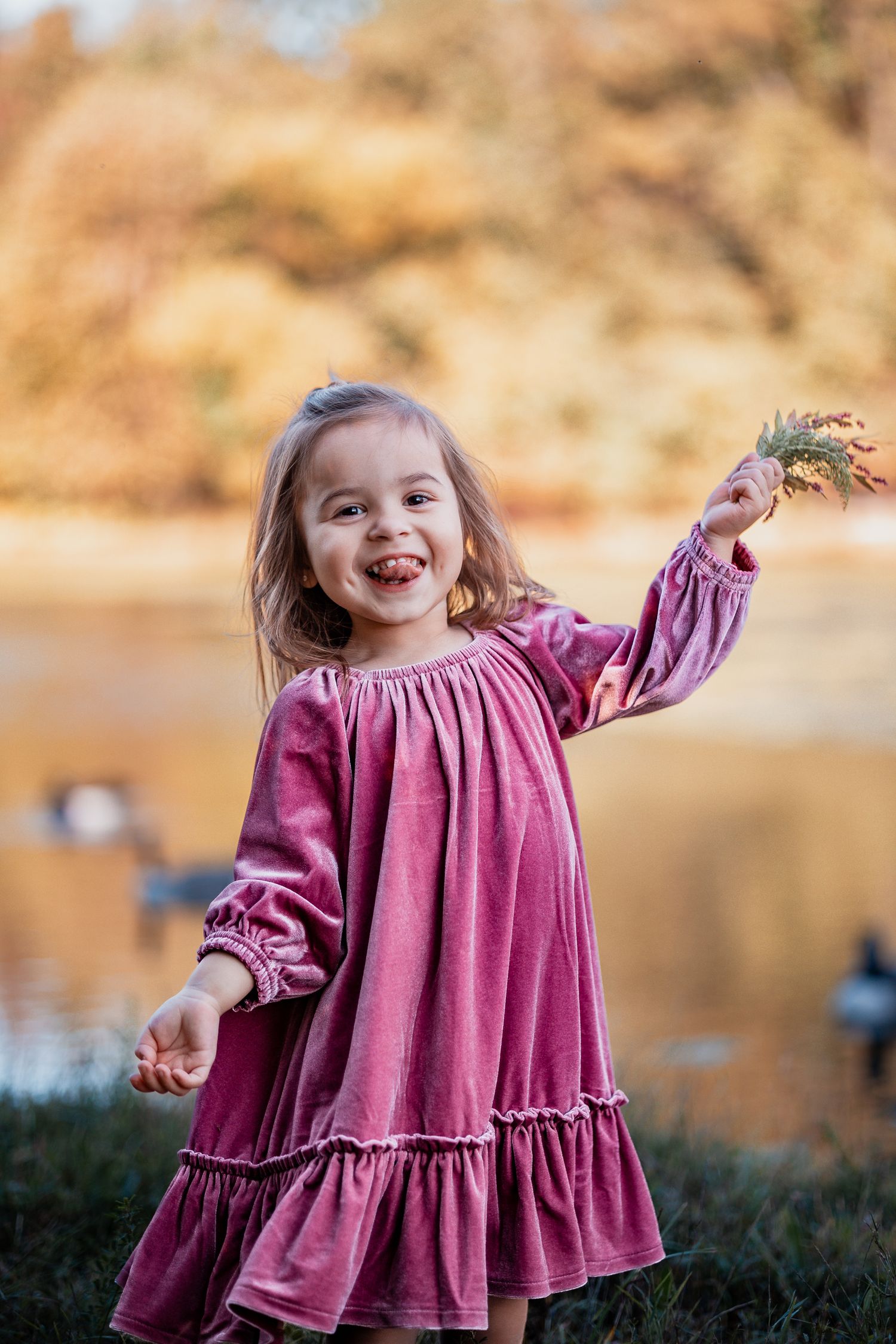 A child in a velvet pink dress plays joyfully by a lake during autumn with golden foliage in the background.