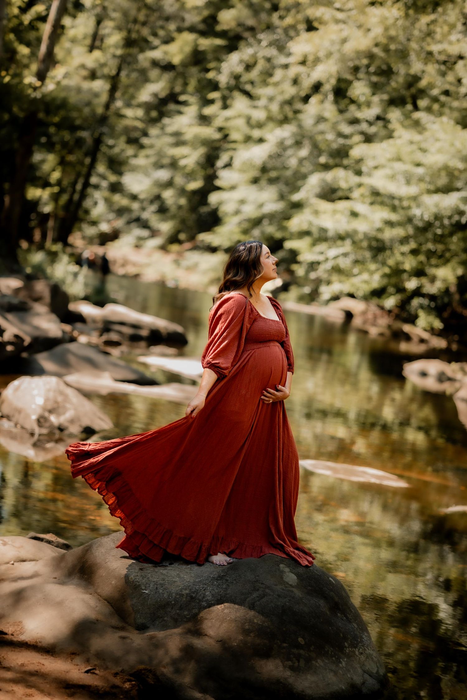 Pregnant woman in flowing red dress standing on rocks by a serene forest creek, cradling her baby bump.