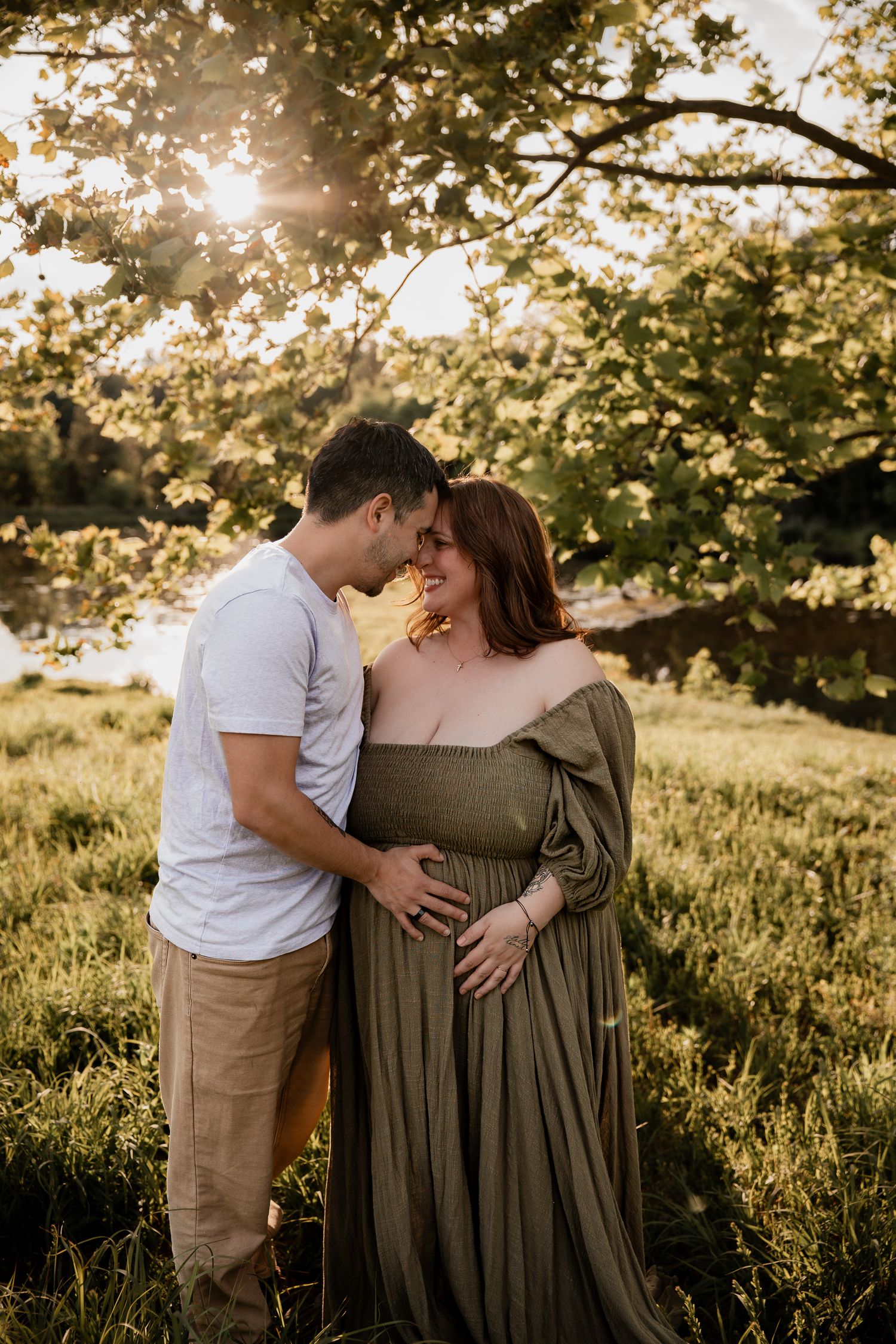 Couple sharing a tender maternity moment outdoors at golden hour, woman in olive green dress cradling baby bump under sunlit trees.