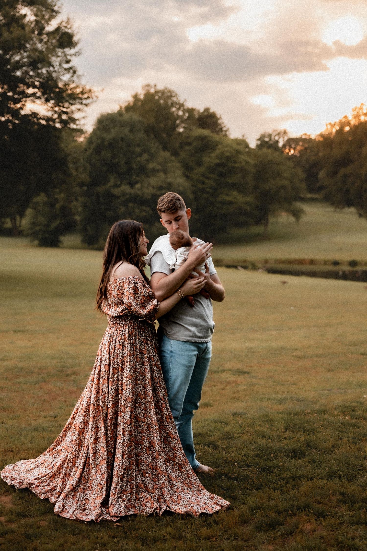 A couple shares a tender moment at golden hour, the woman in a floral maxi dress as the man kisses their newborn baby.
