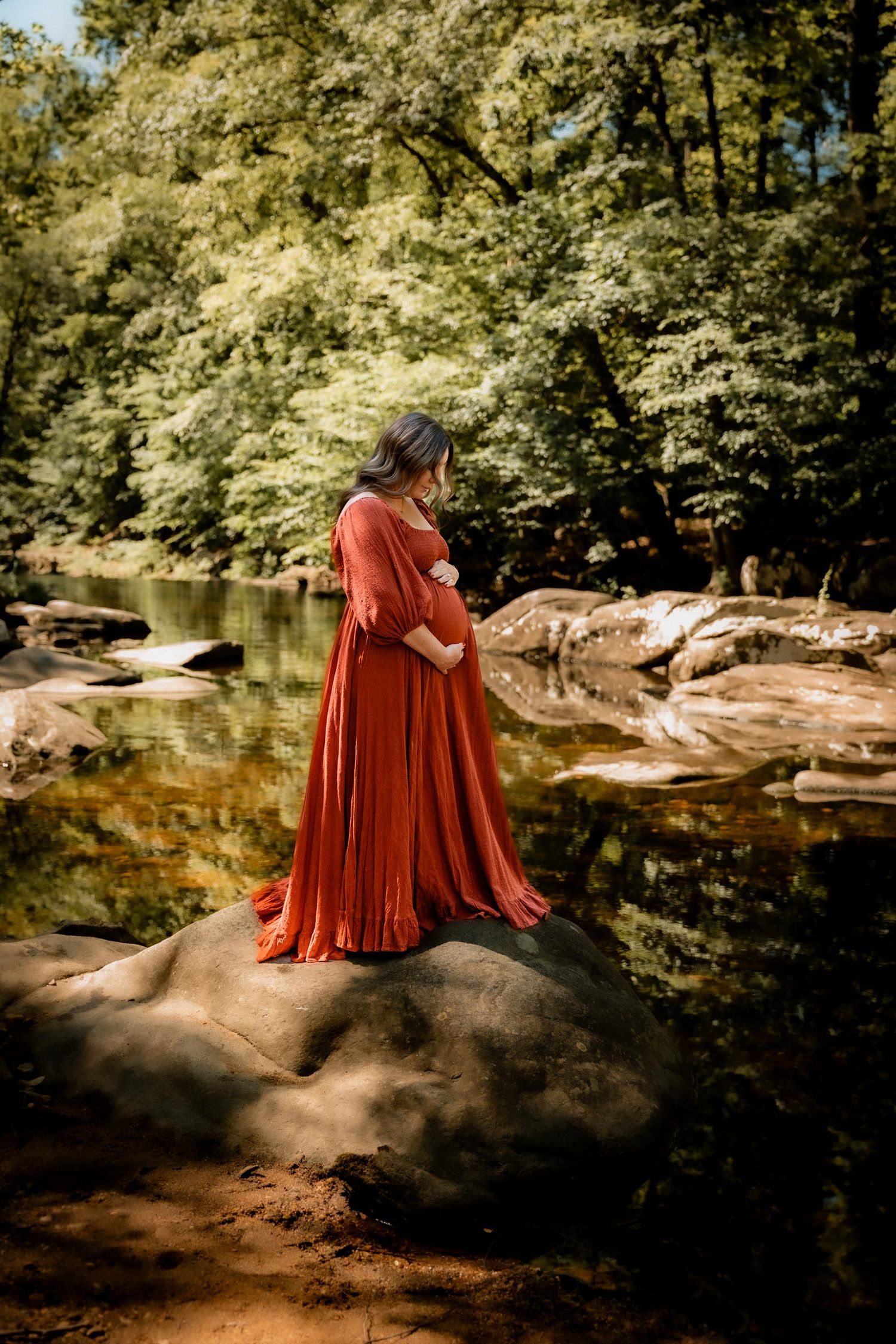 Pregnant woman in flowing rust dress standing on rocks by a serene forest river, cradling her baby bump.