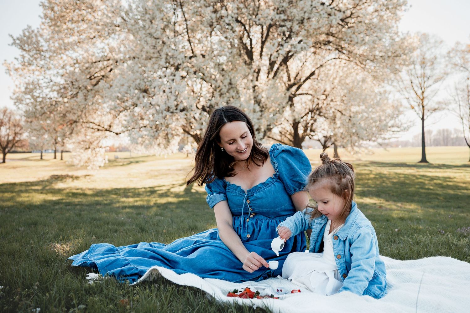 A mother in a blue dress sits on a white blanket with her young daughter under blooming trees in a park.