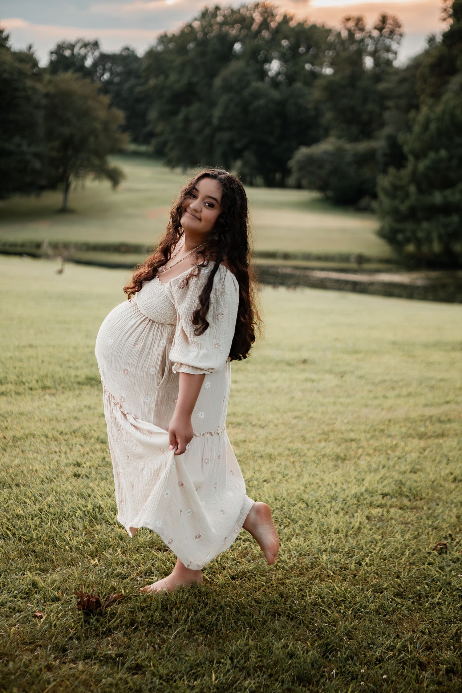 Woman in white dress spinning playfully in grassy field at sunset while surrounded by oak trees.