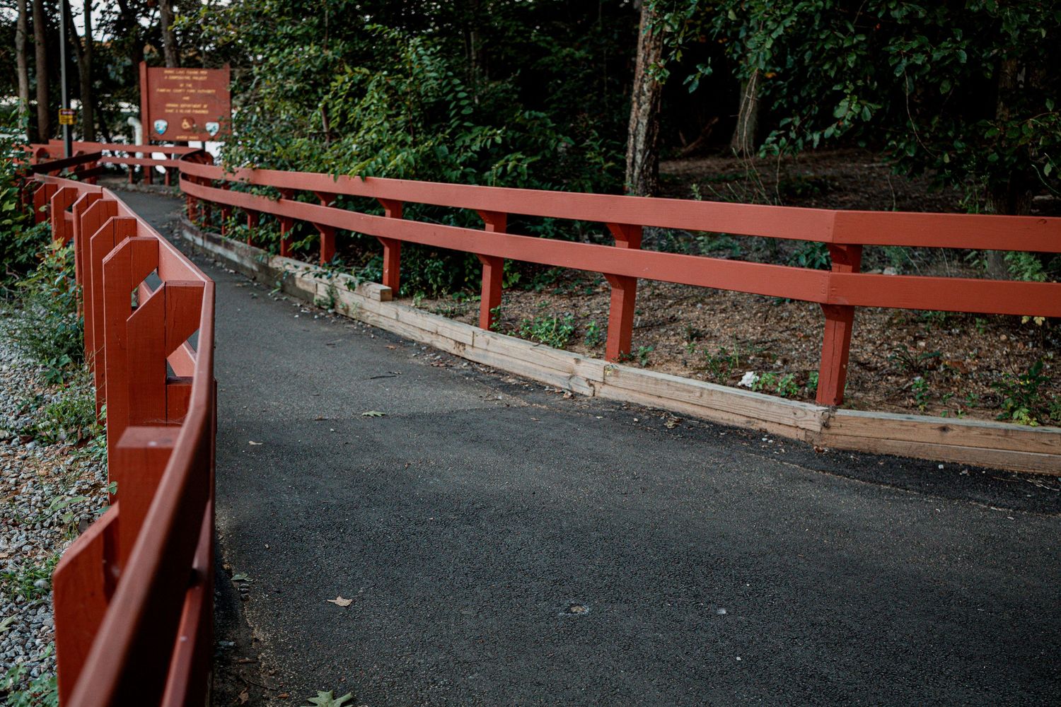 A winding paved path bordered by red metal railings leads through a lush green wooded area.