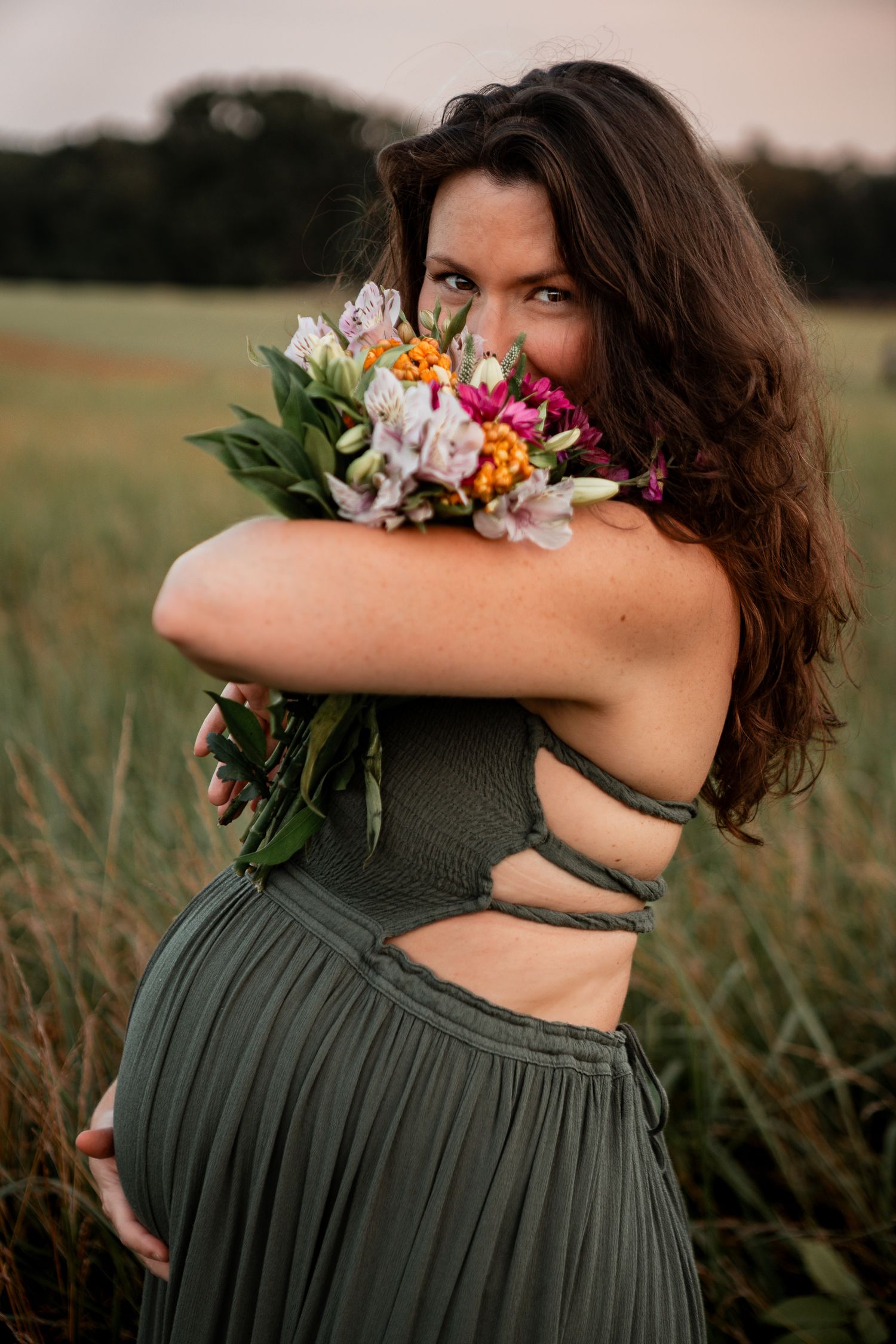 A woman in a sage green dress holds a colorful bouquet of flowers in front of her face while standing in a field.