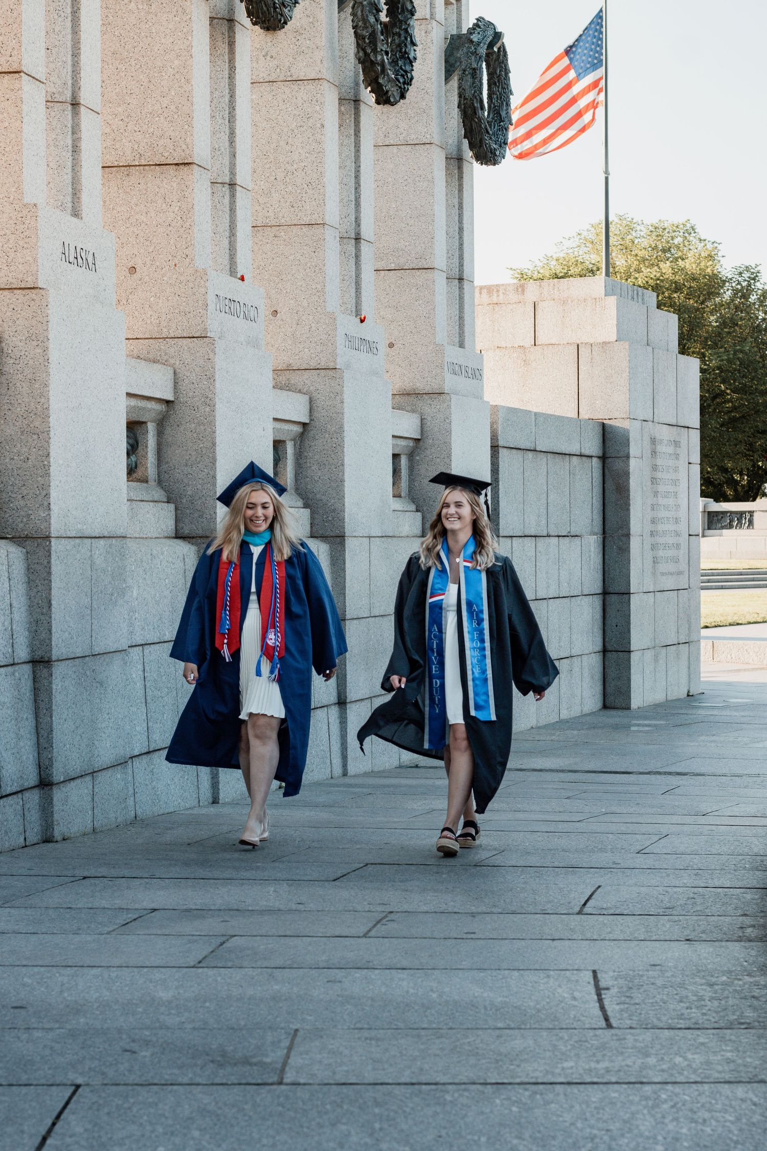 Graduates walk together in caps and gowns near stone columns with an American flag waving in the background.