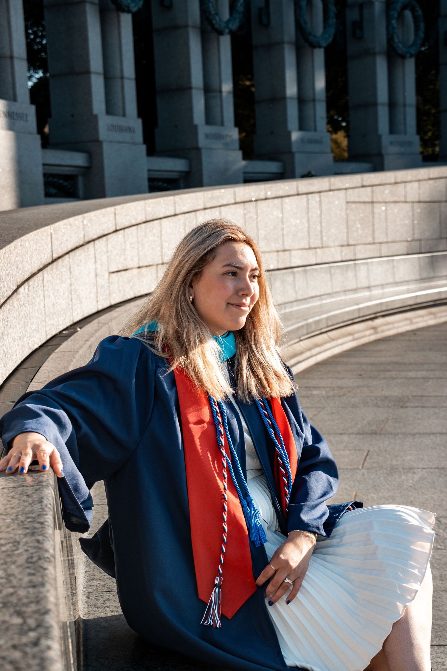 Graduate posing in academic regalia with blue robe and red stole against architectural columns on sunny day.