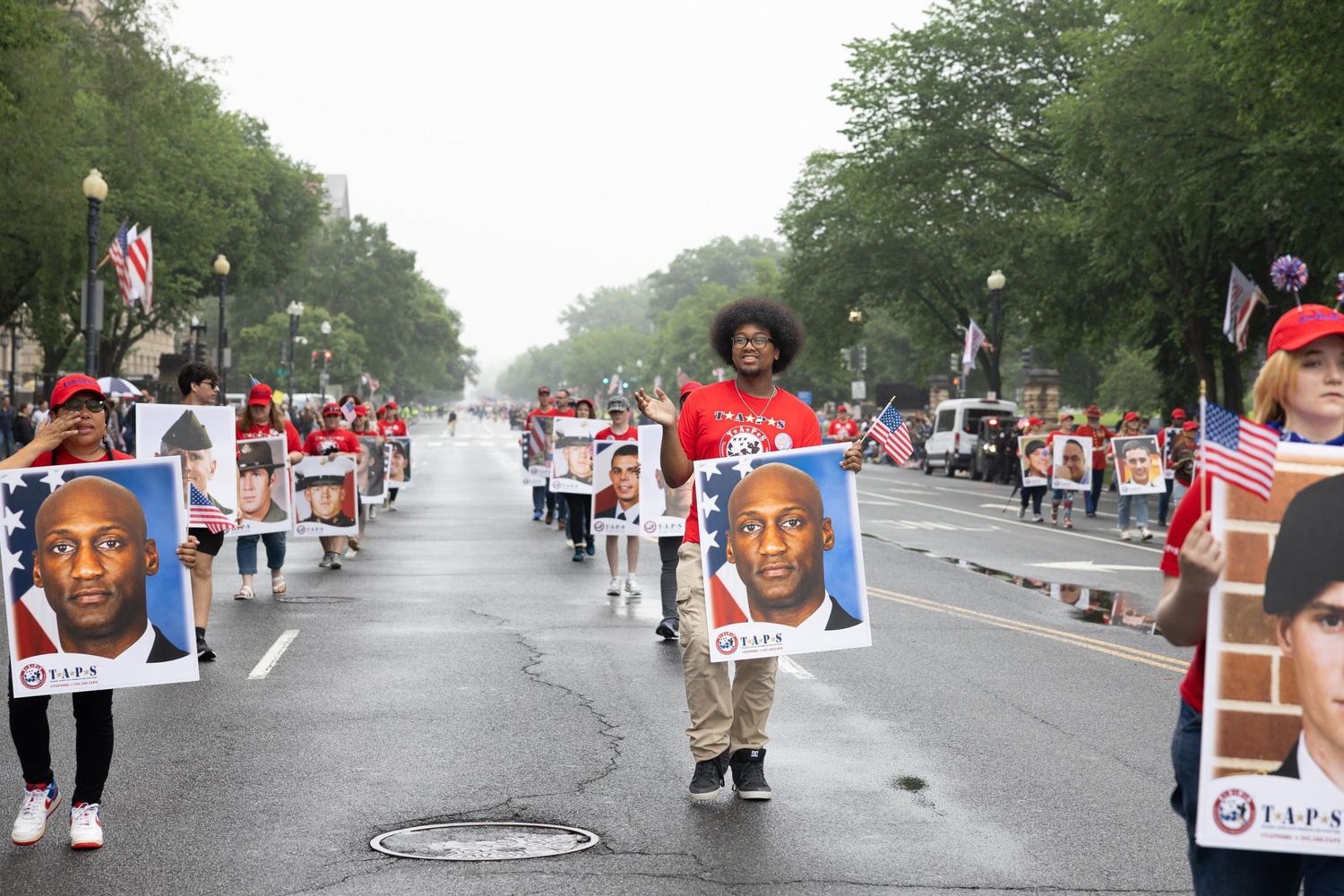 People marching in a parade holding large portrait signs and American flags on a wide urban street.