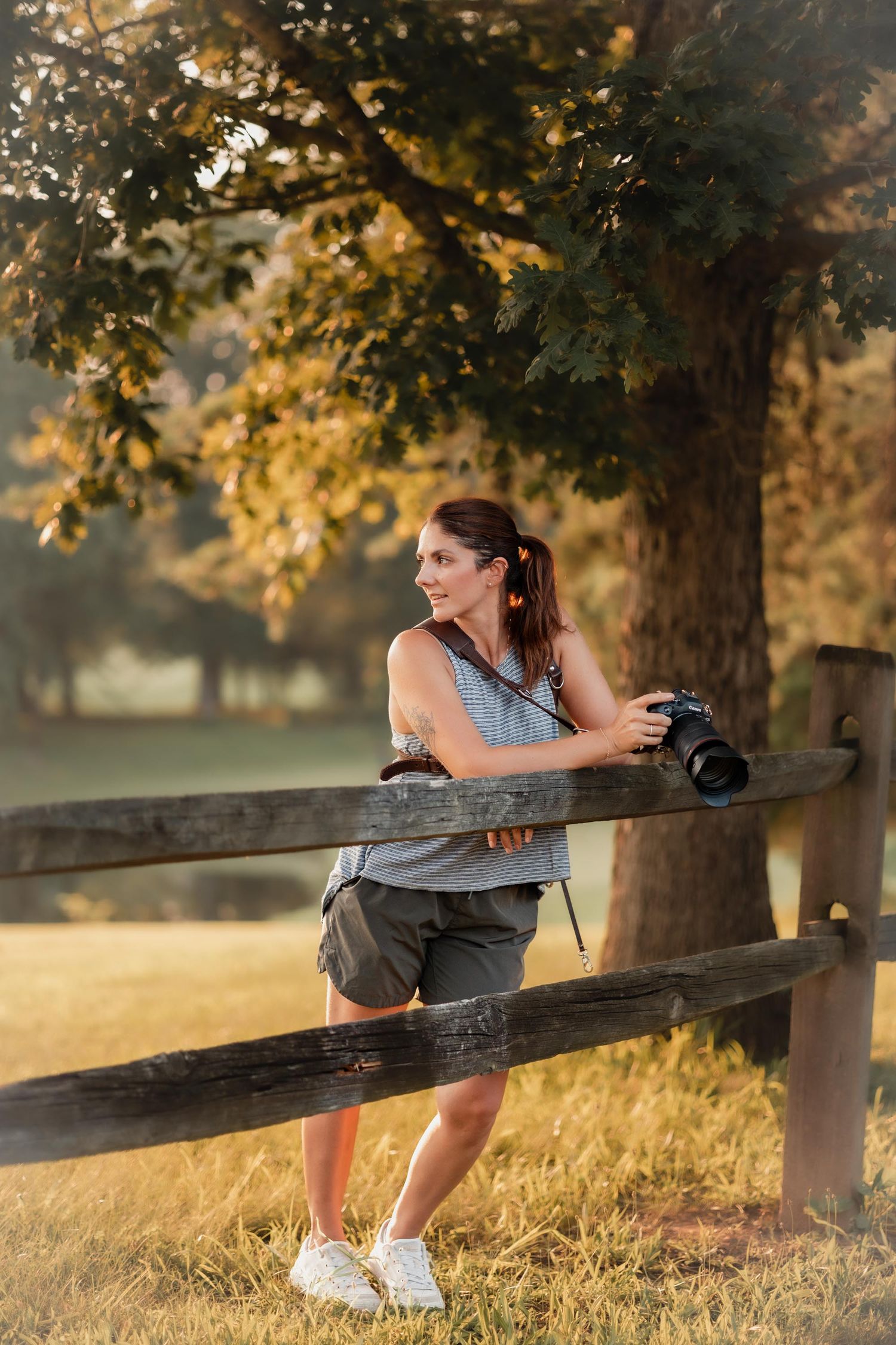 Woman photographer leaning on wooden fence holding DSLR camera in golden hour sunlight with trees in background.