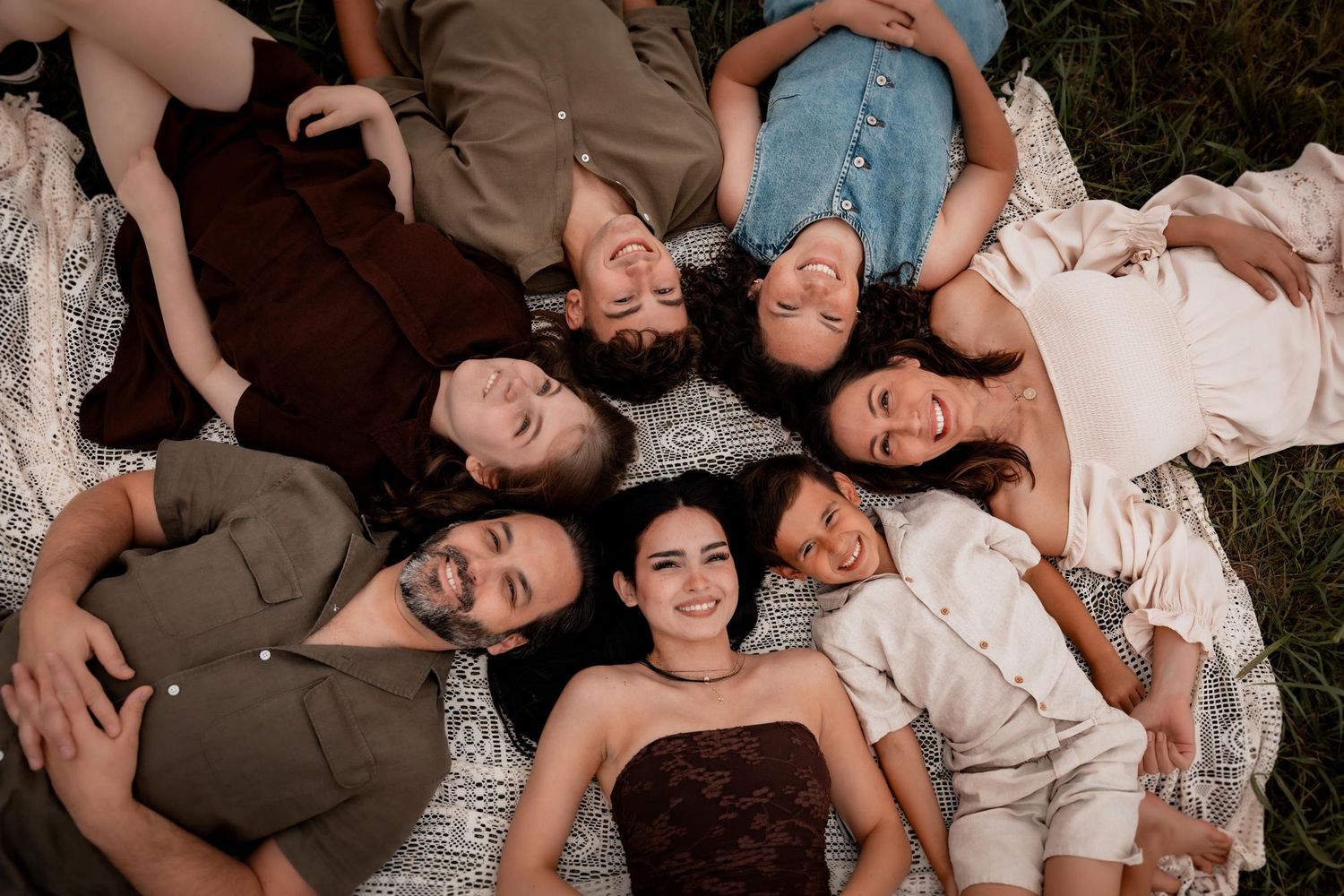 Seven people lying in a circle on a blanket outdoors, smiling up at the camera in a joyful family portrait.