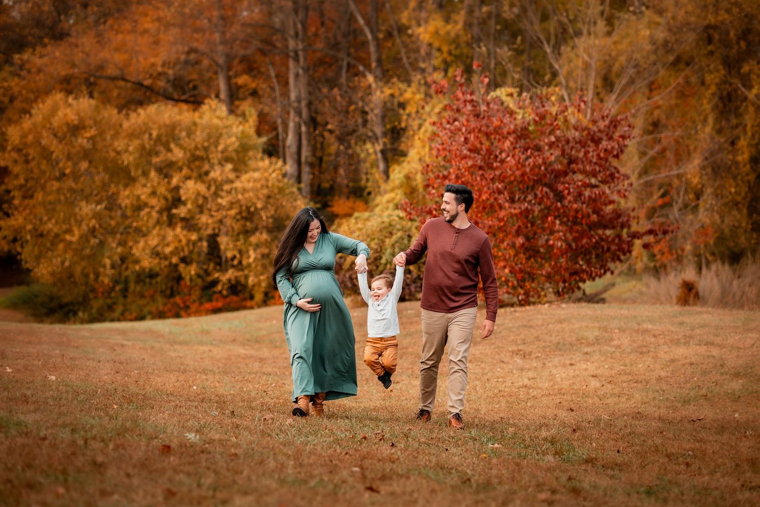 Pregnant family walking outdoors in autumn park, holding toddler's hands among vibrant fall foliage.