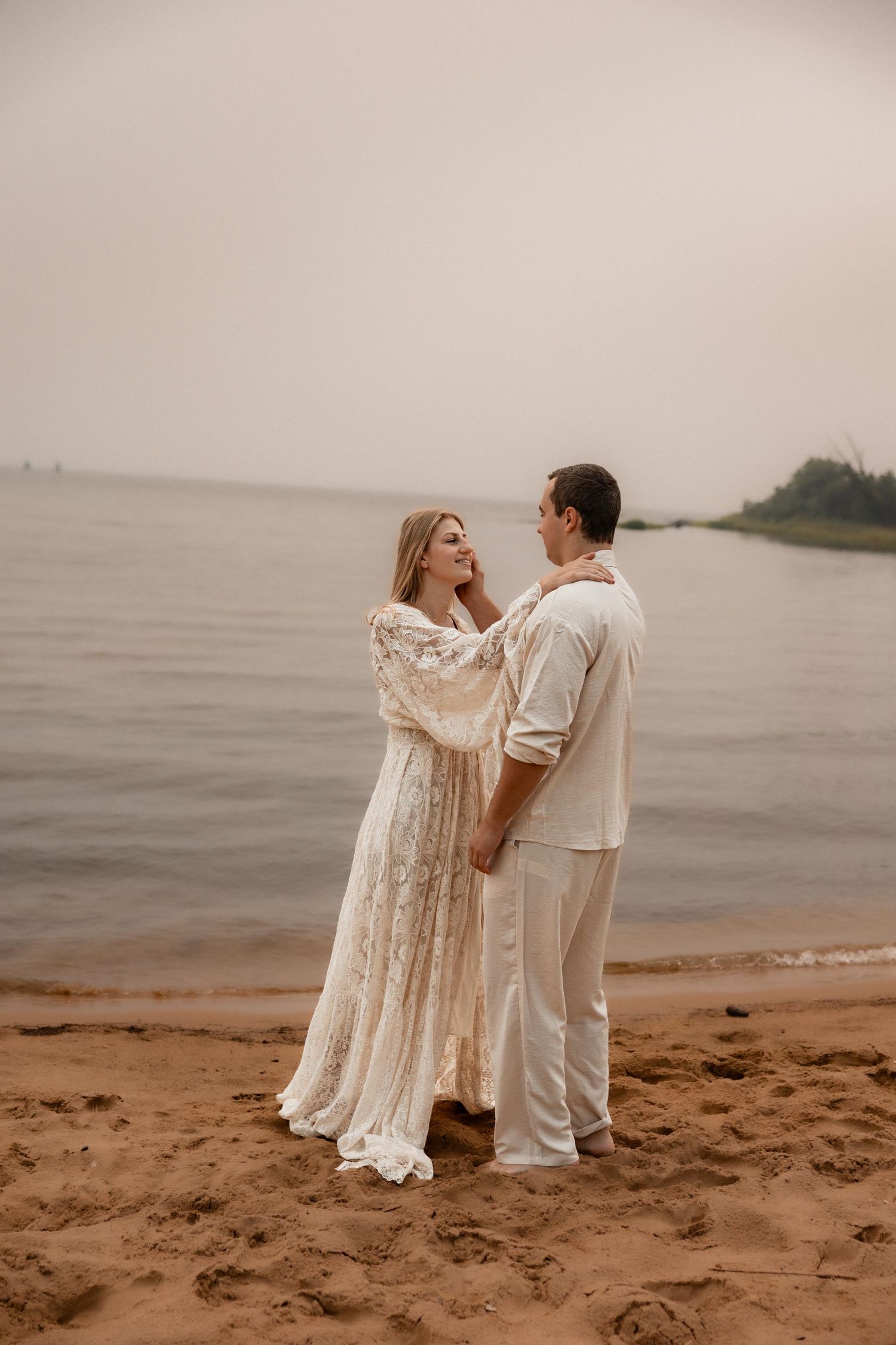 Romantic couple in white attire standing on sandy beach by calm water, woman in lace dress touching man's face.