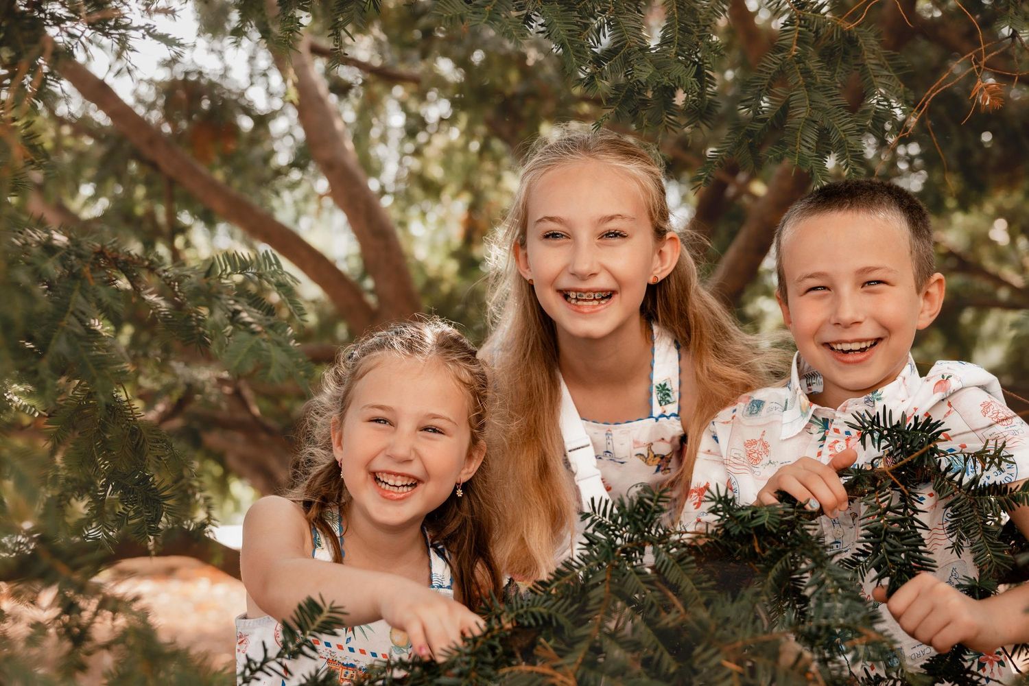 Three happy children smiling outdoors among green tree branches in a warm, sunny setting.
