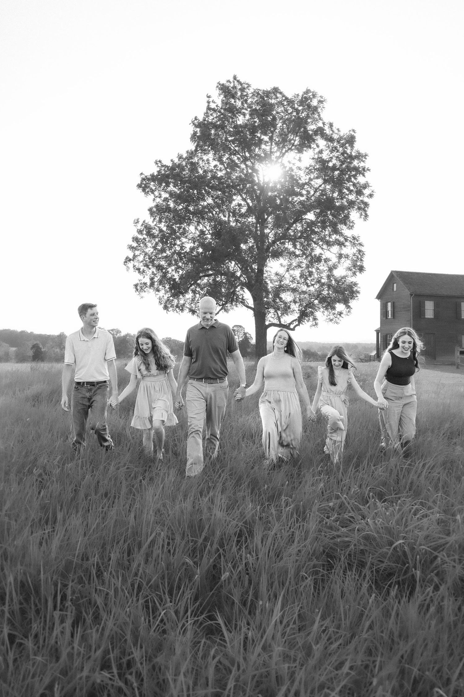 Black and white photo of a family of six holding hands walking through a field with a tree and barn in the background.