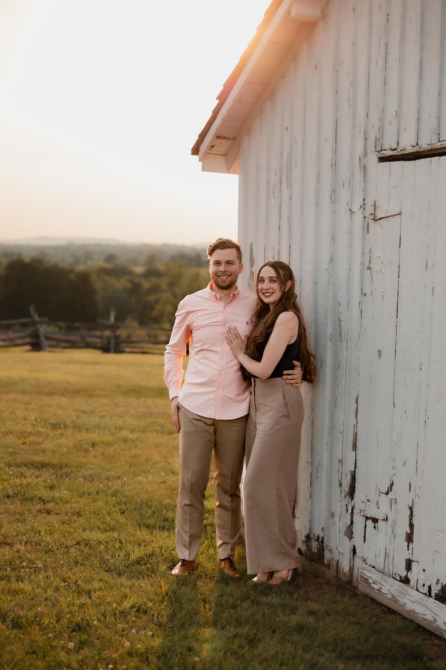 A young couple leans against a white barn wall smiling at sunset on a green countryside.