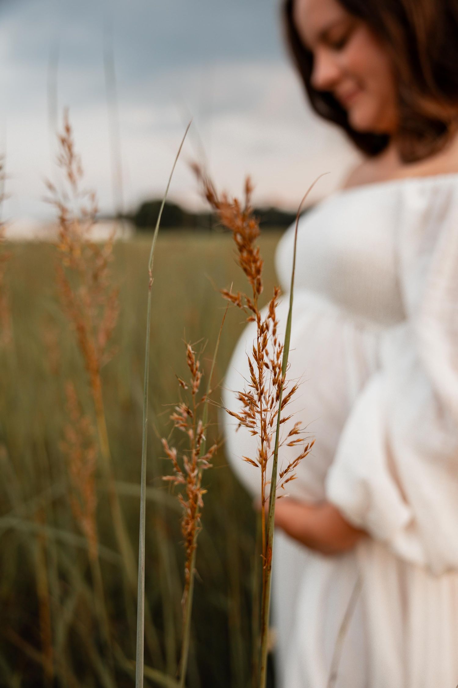 Pregnant woman in white dress standing in a green field with tall golden grass in the foreground.