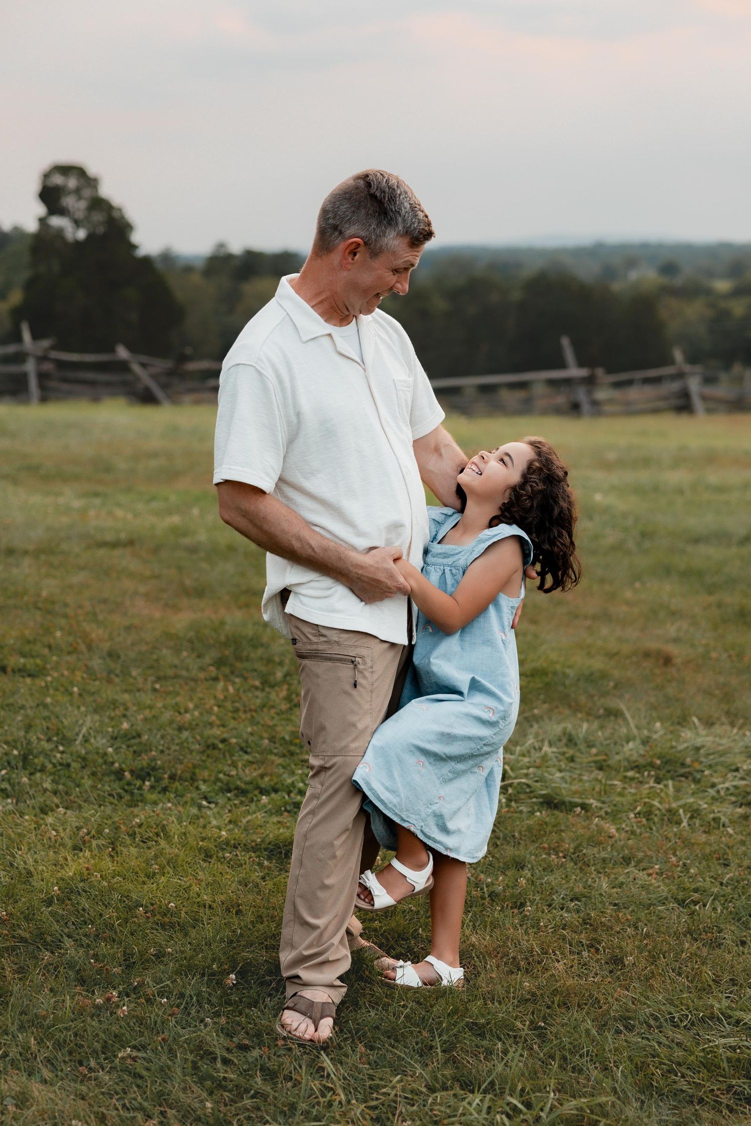 A young curly-haired girl in a blue dress laughs and dances with her father in a grassy meadow.
