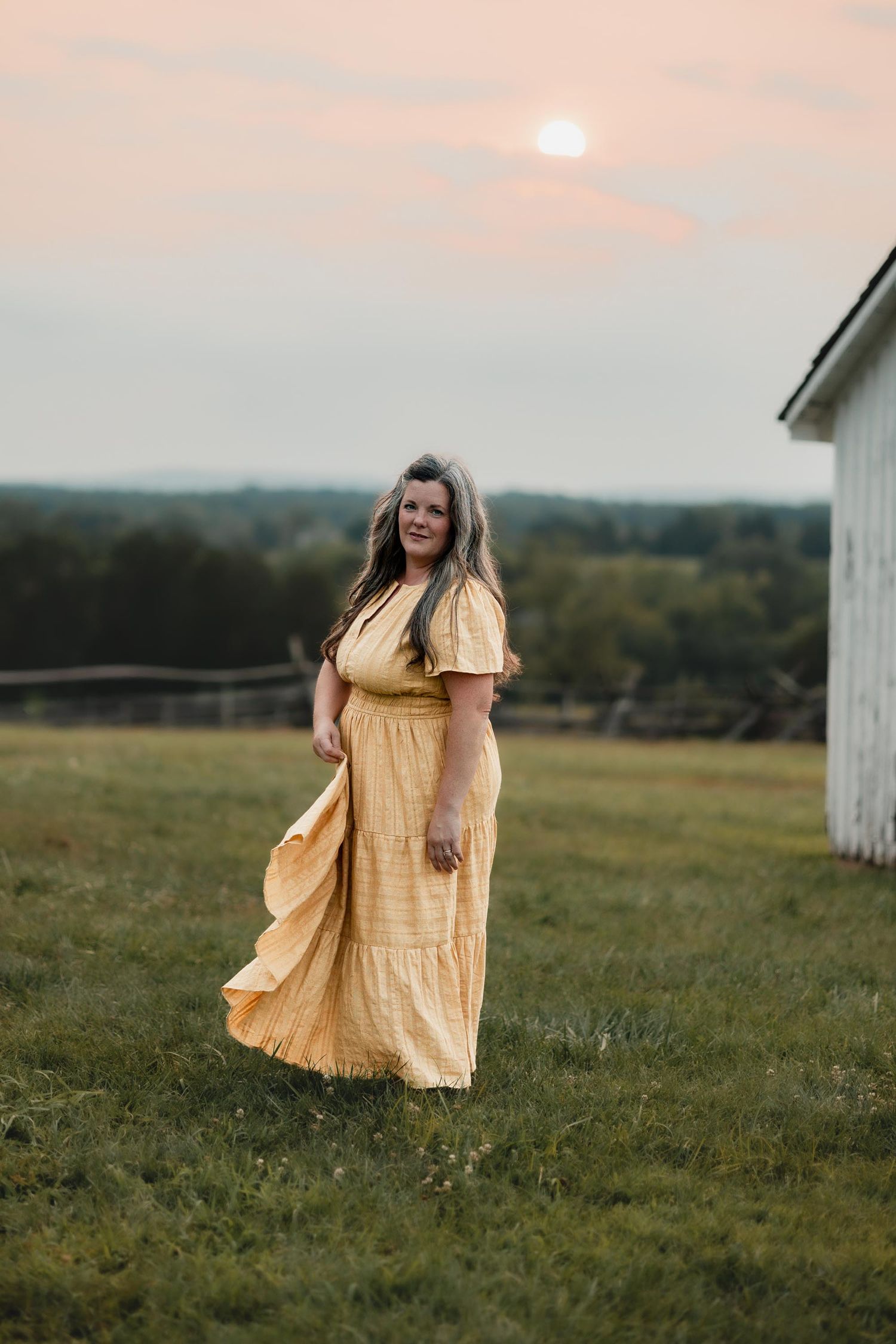 A woman in a flowing yellow maxi dress stands in a green field at sunset near a white barn.