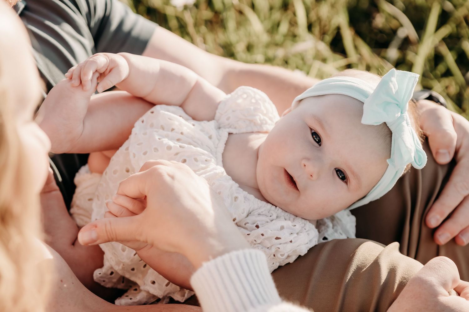 A baby wearing a mint green headbow and white lace dress rests in someone's arms outdoors.
