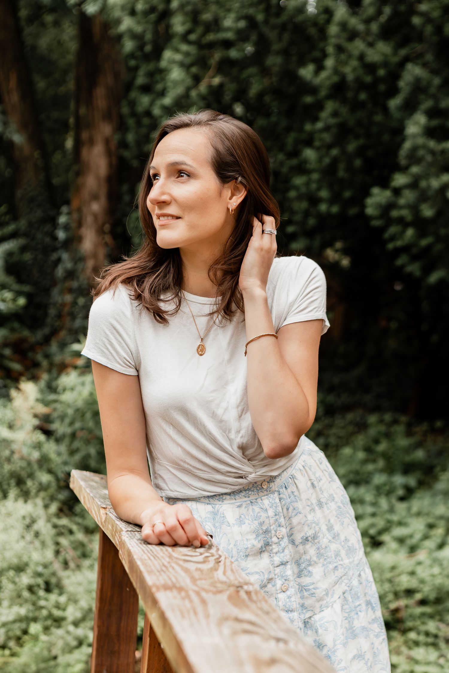A person in a white t-shirt and floral skirt leans on a wooden railing in a dreamy forest setting.