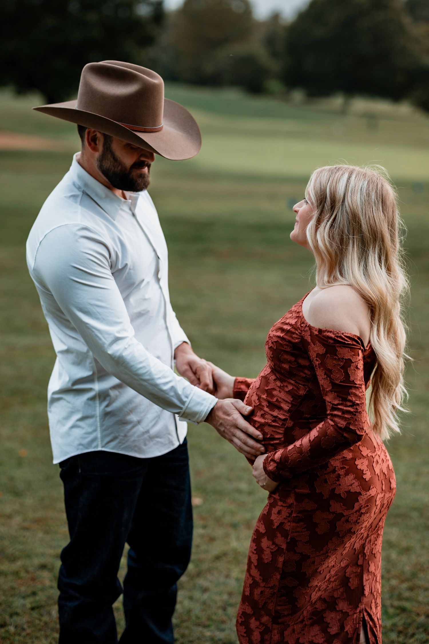 Romantic maternity photos in a park with couple in rust dress and western attire.