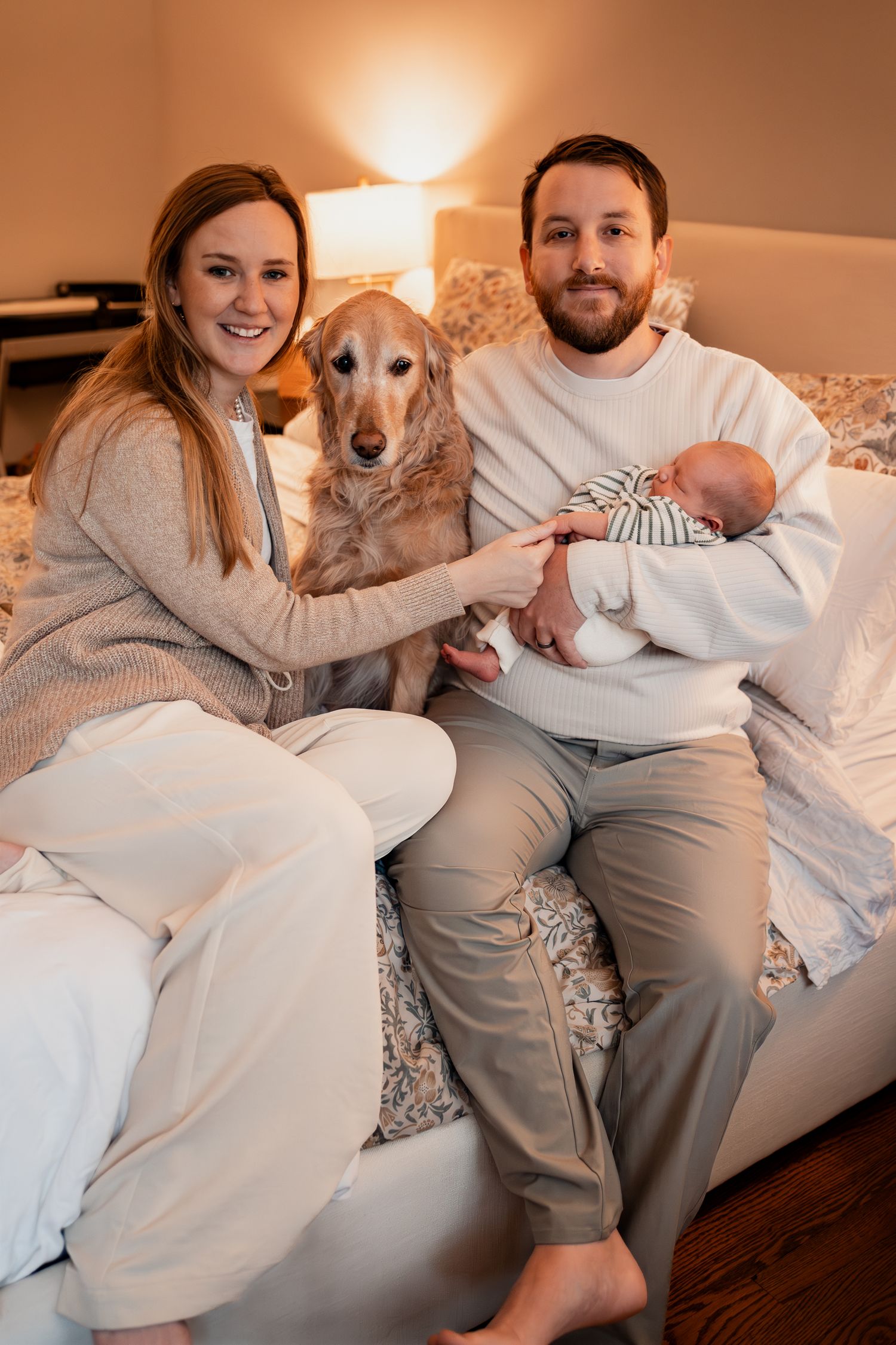 A cozy family moment on a bed with their golden retriever and newborn baby in a warmly lit room.