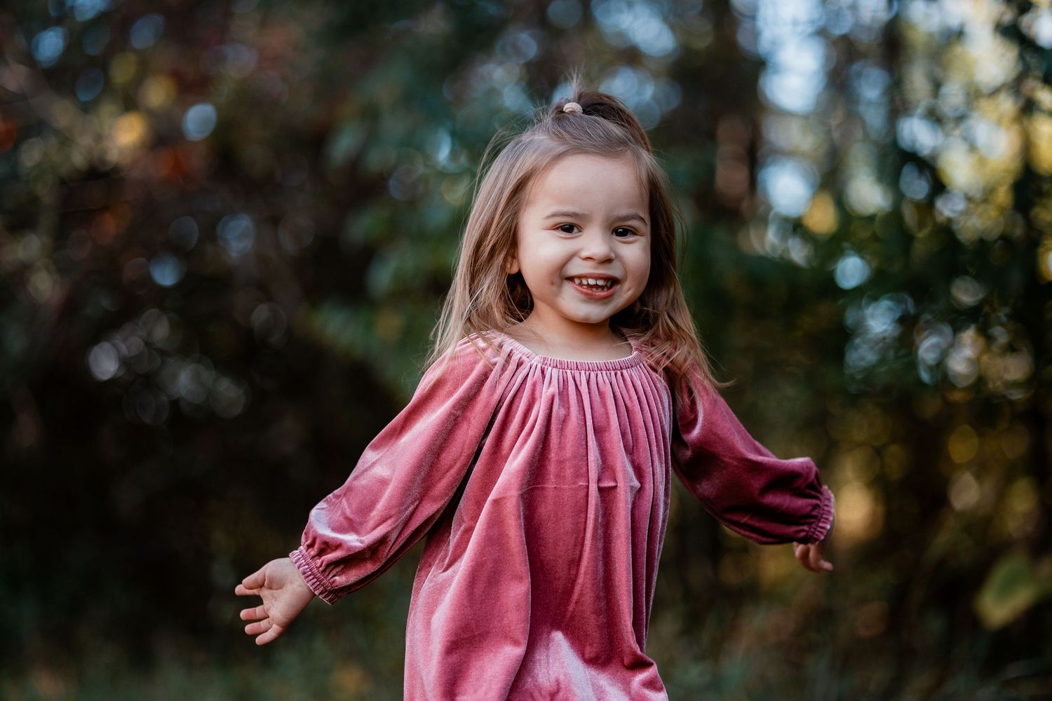 A child in a pink dress twirls playfully in a sunlit outdoor setting with autumn trees in the background.