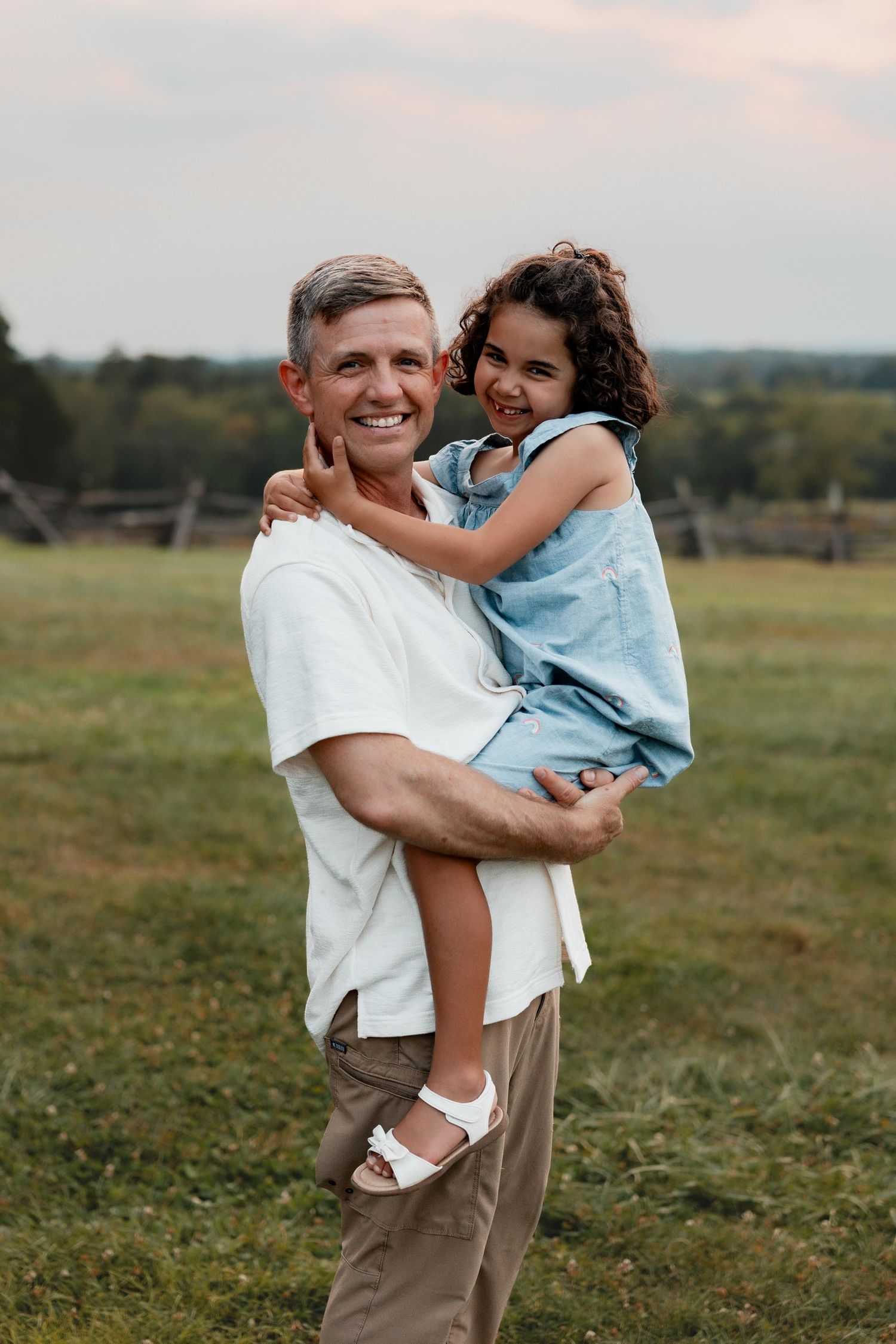 A smiling man holding a happy young curly-haired girl in a blue dress in an outdoor field setting.