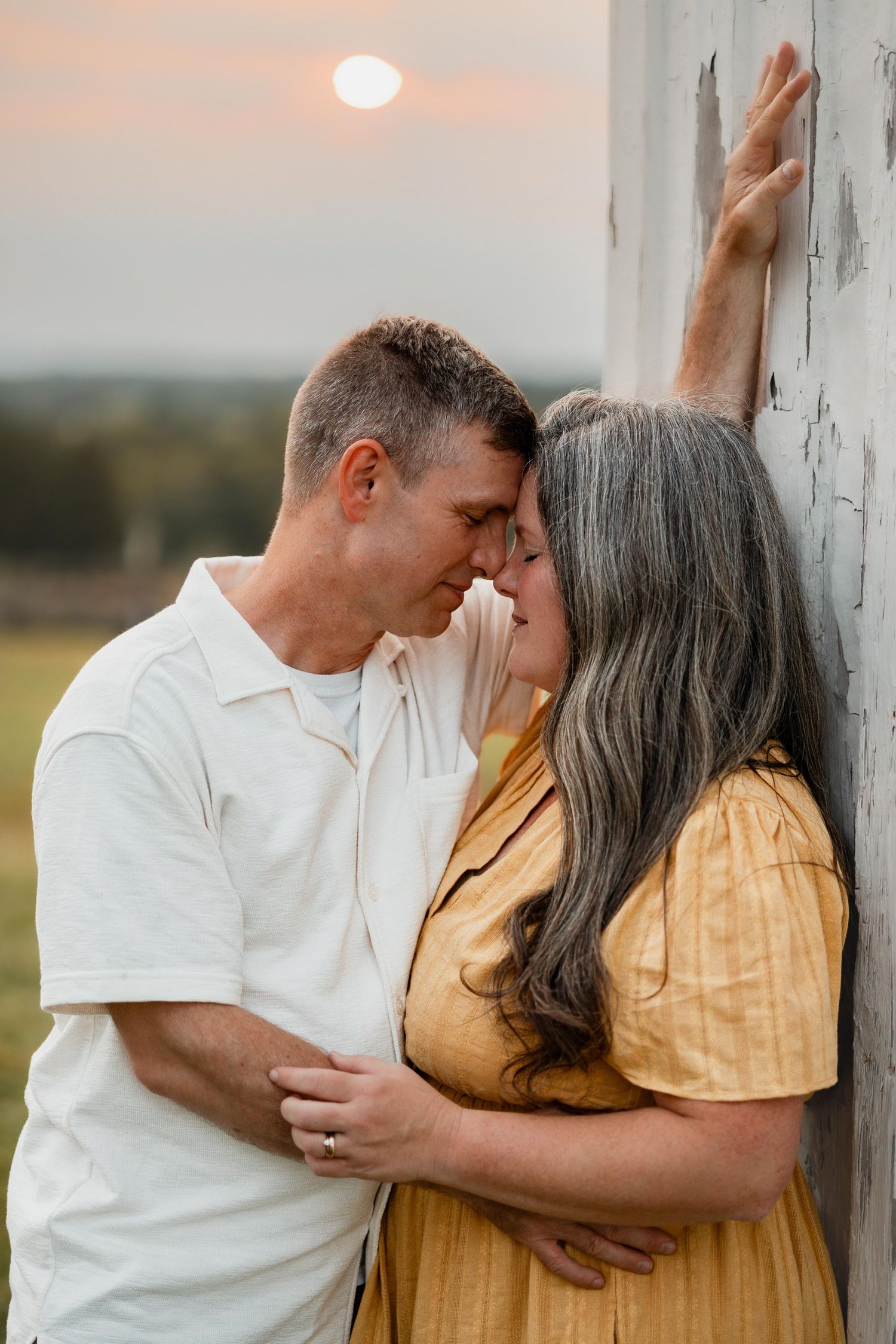 A romantic couple leans in for a kiss against a weathered white barn wall at sunset.