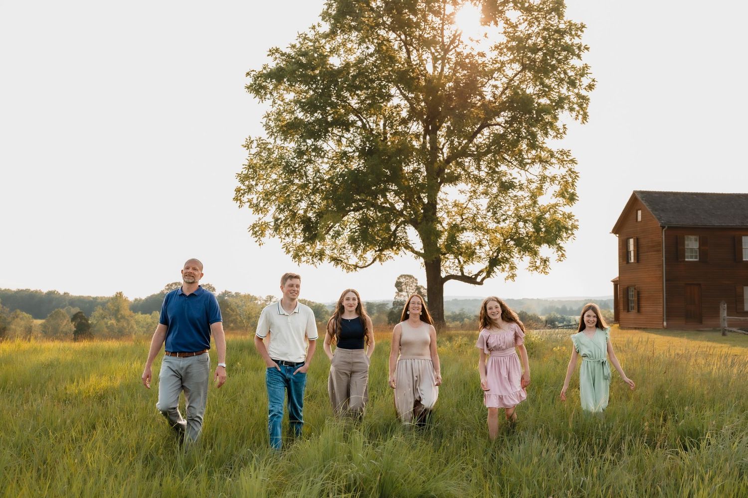 Family of six walks through a sunlit green field with a large tree and red barn in the background.