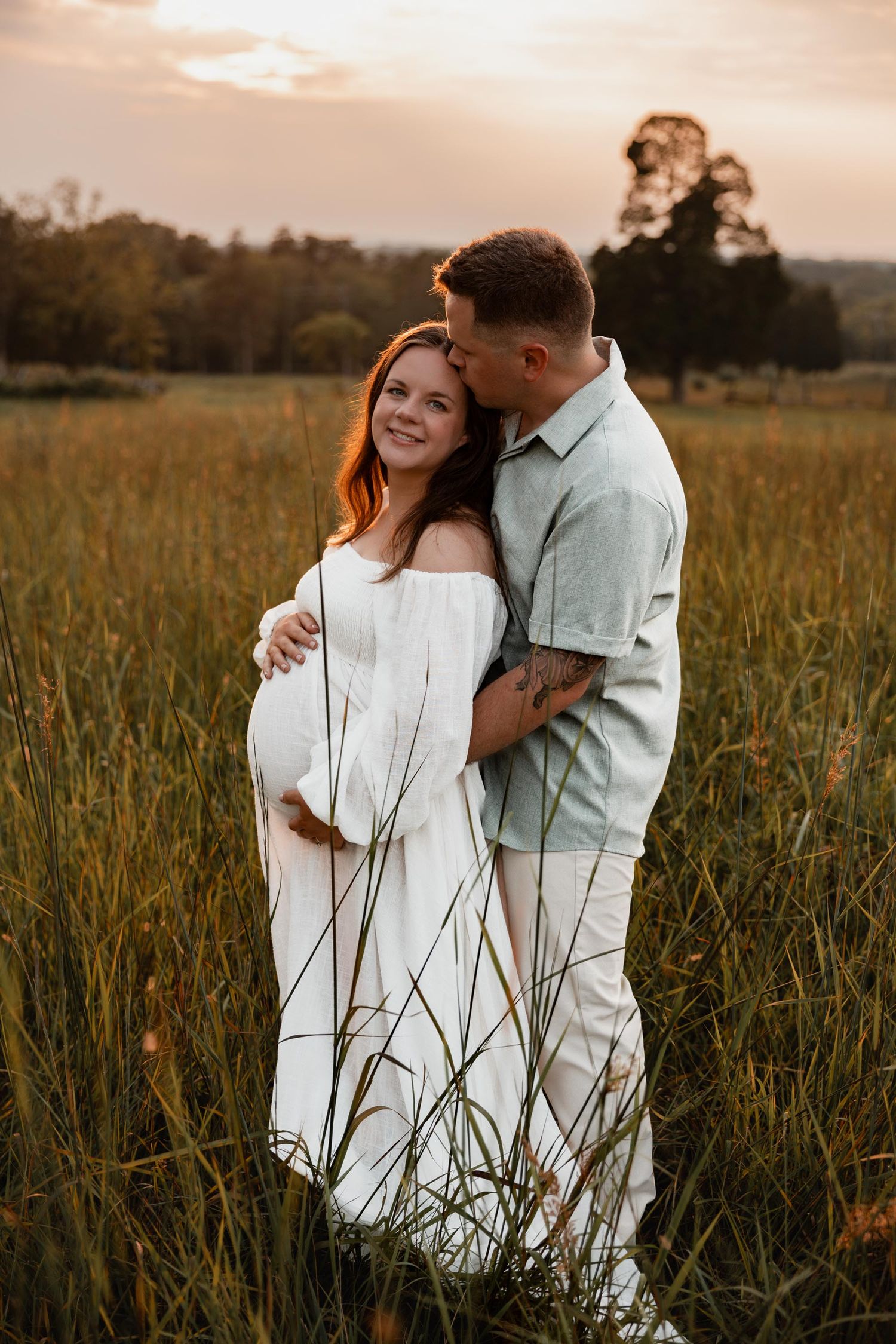 Couple embracing in golden field at sunset, pregnant woman in white off-shoulder dress smiling joyfully.