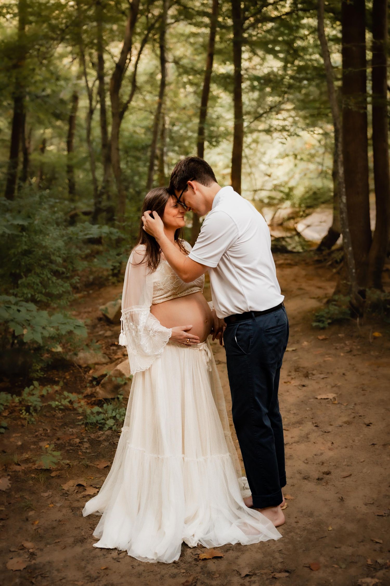 Expecting couple sharing tender forehead touch on forest path, woman in flowing white maternity gown cradling baby bump.