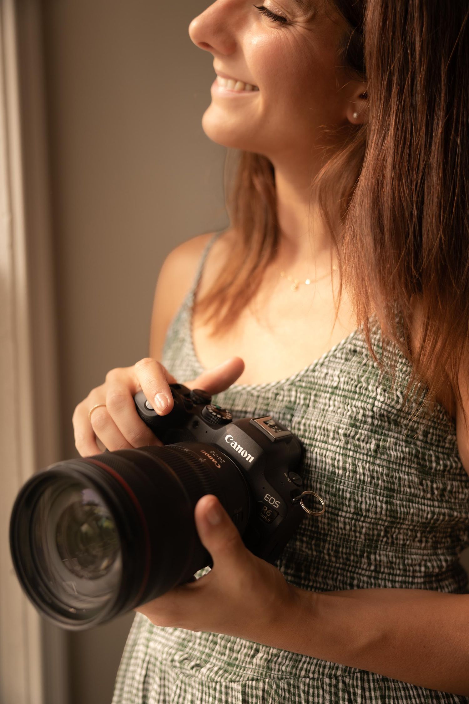 Smiling woman holding a Canon EOS DSLR camera with a large lens, wearing a green dress near a window.