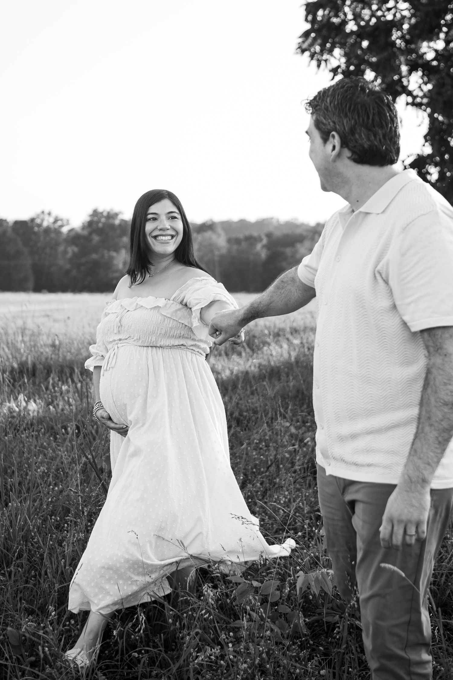 A couple shares a joyful moment in a field during maternity photoshoot, with her white dress flowing in the breeze.